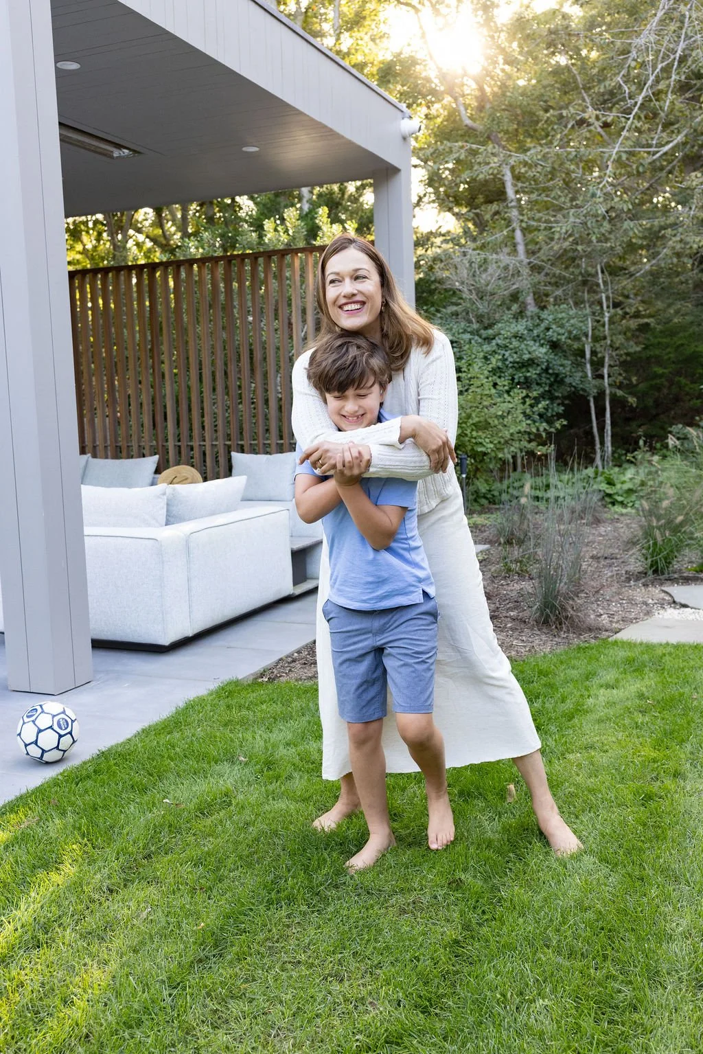 A woman and a boy playing outdoors on a grassy yard, embracing and smiling as sunlight filters through trees in the background.