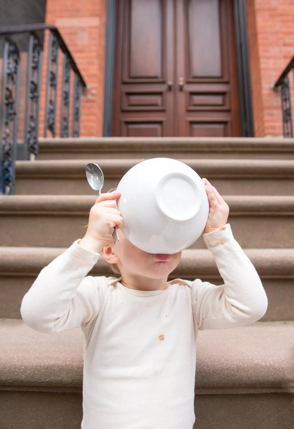 A young child standing on outdoor stone steps, holding a large white bowl over their face like a helmet, with a spoon in one hand, in front of a brick building with a wooden door.