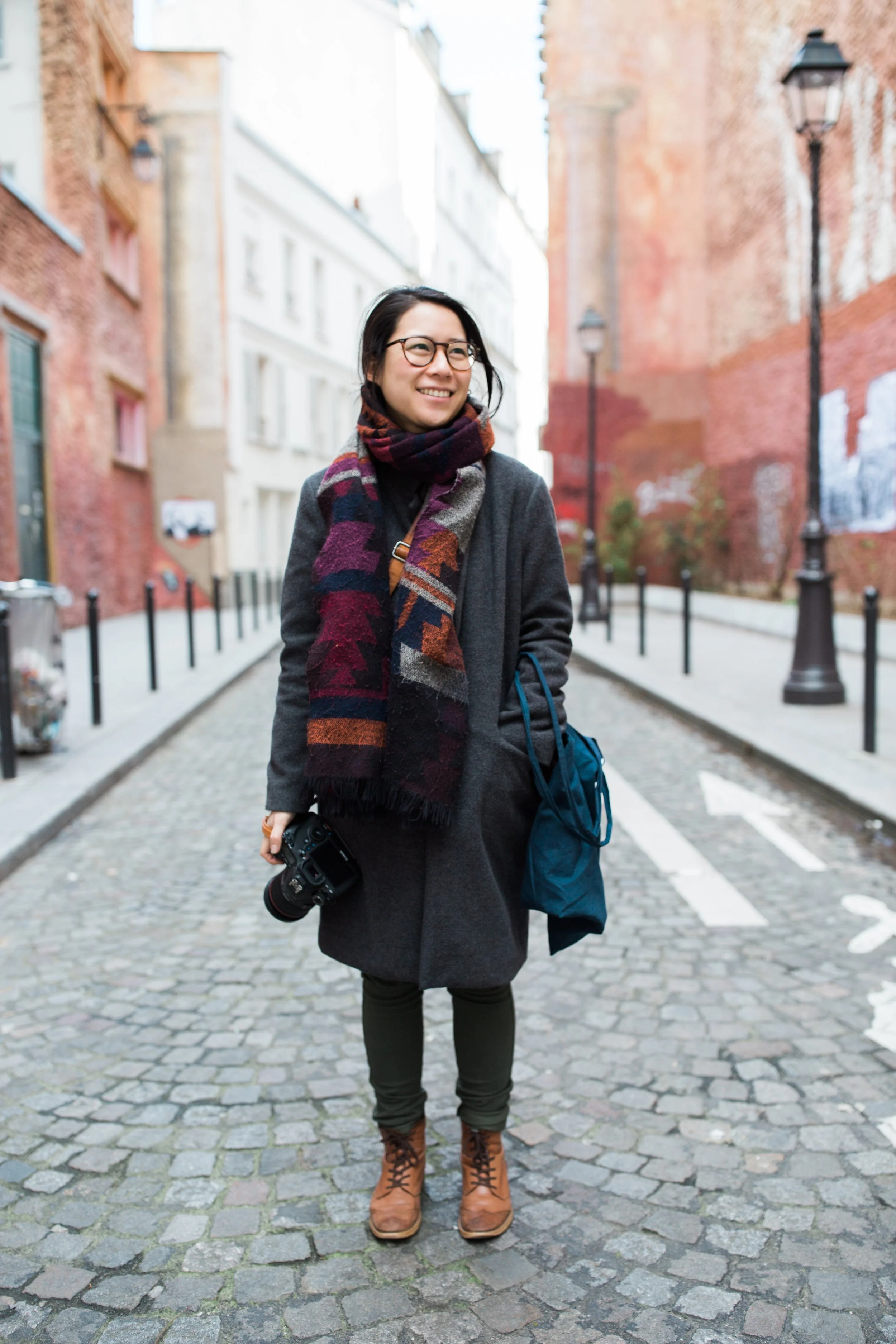 A woman with glasses and a multicolored scarf smiling while standing on a cobblestone street holding a camera with a strap, with old brick buildings and streetlights in the background.