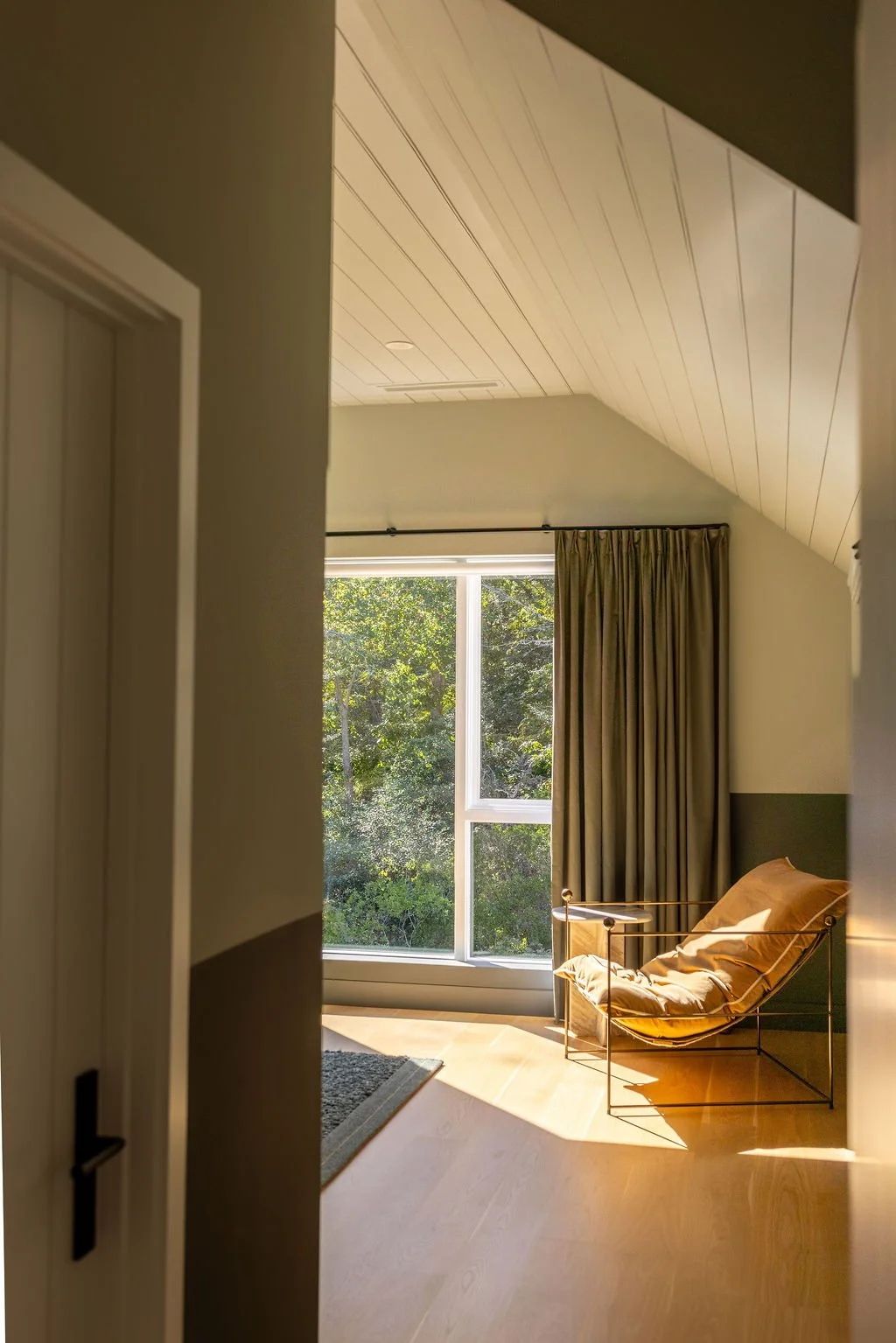 Room with a large window showing green trees outside, a beige recliner chair with pillows, beige curtains, wood flooring, and an angled white ceiling.