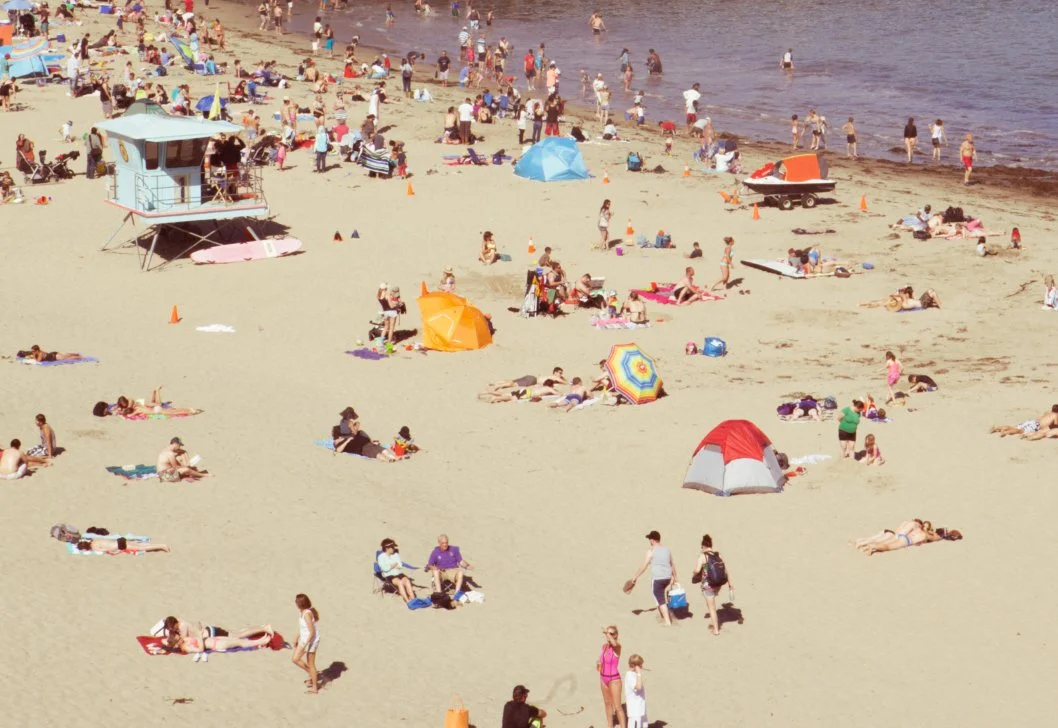 Crowded beach with beachgoers sunbathing, swimming, and walking, along with colorful umbrellas, tents, and a lifeguard tower.