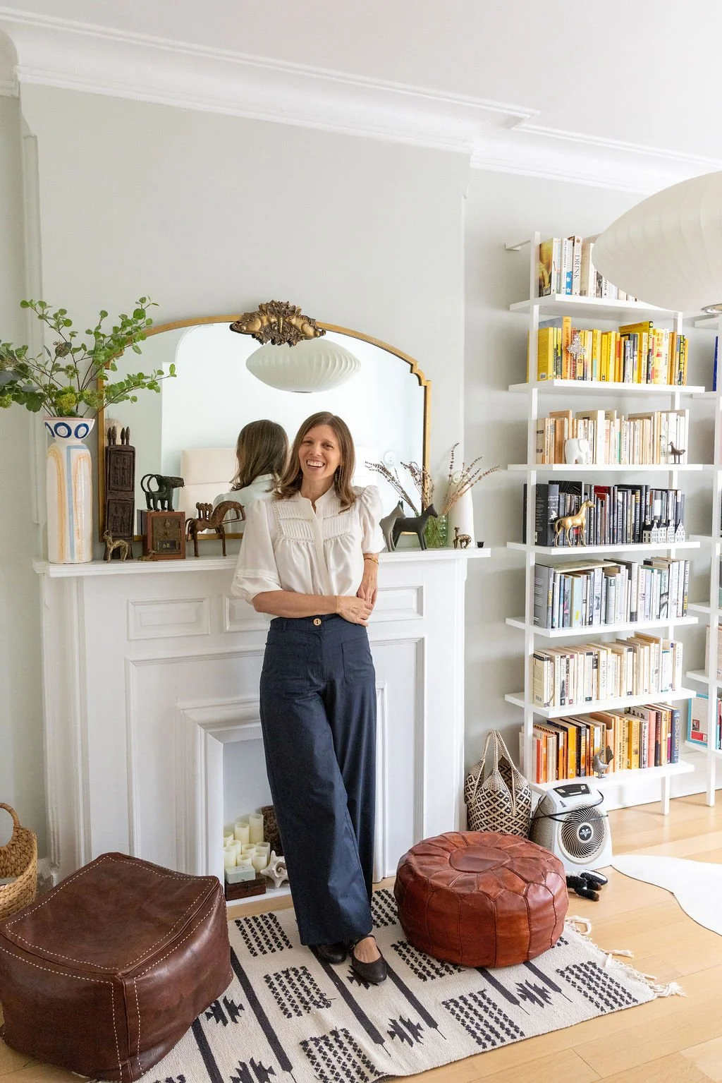 A woman standing in front of a fireplace mantel with a mirror above it, smiling and wearing a white blouse and dark pants, with a bookshelf filled with books and decorative items in the background.