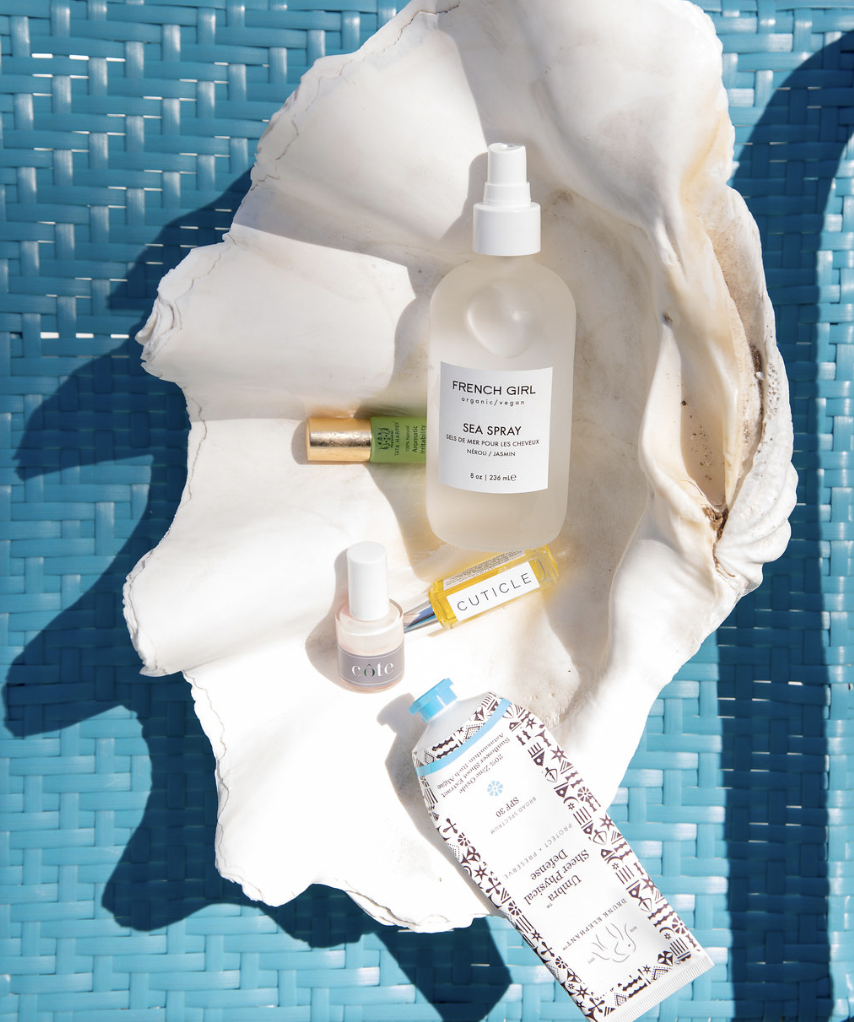 Assorted skincare products and sun protection items on a large seashell sitting on a blue woven surface. The products include a sea spray, lip balm, nail polish, and sunscreen.