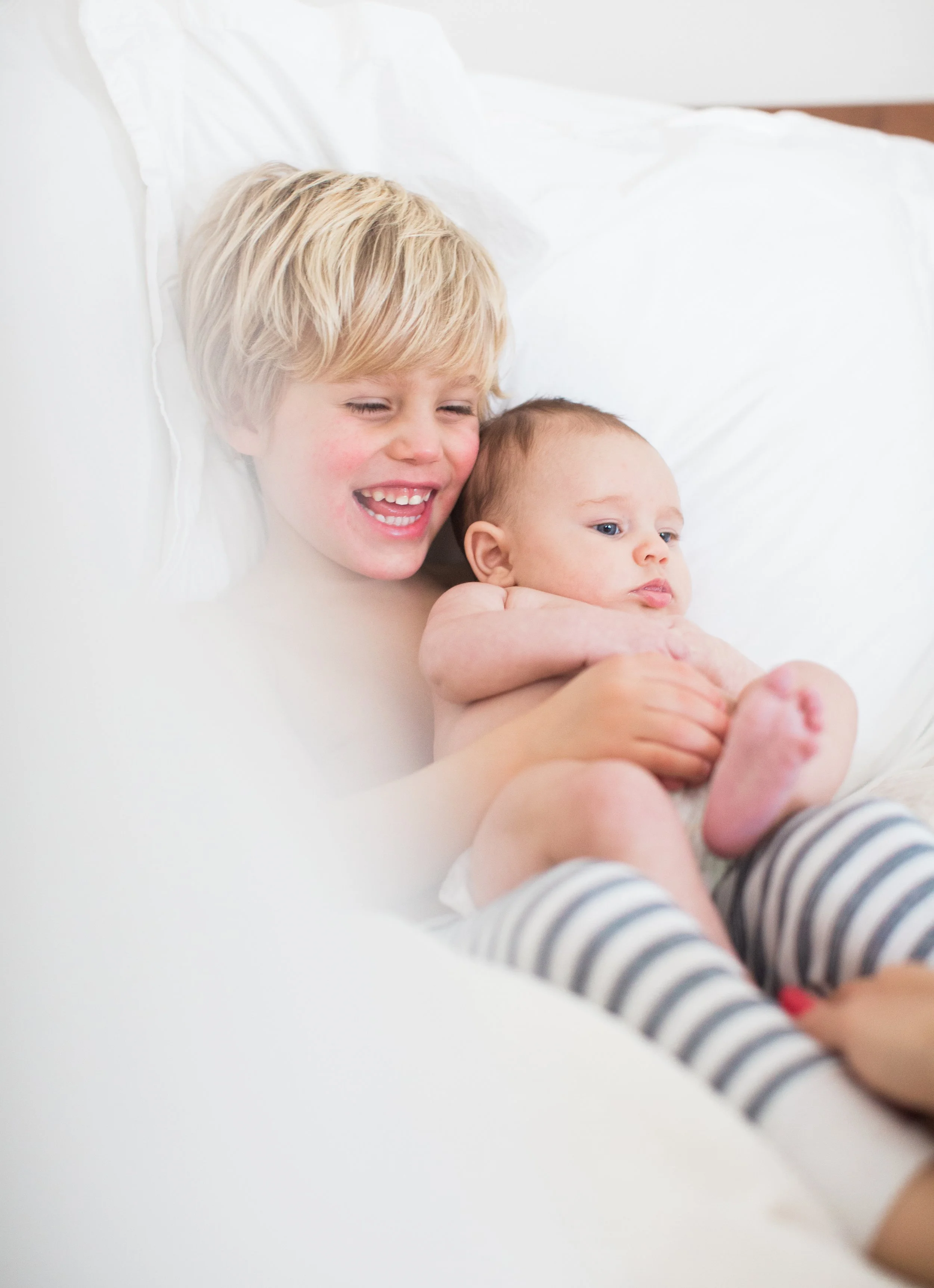 A young boy with blonde hair smiling and laughing while holding an infant in a crib, wearing striped pajamas.
