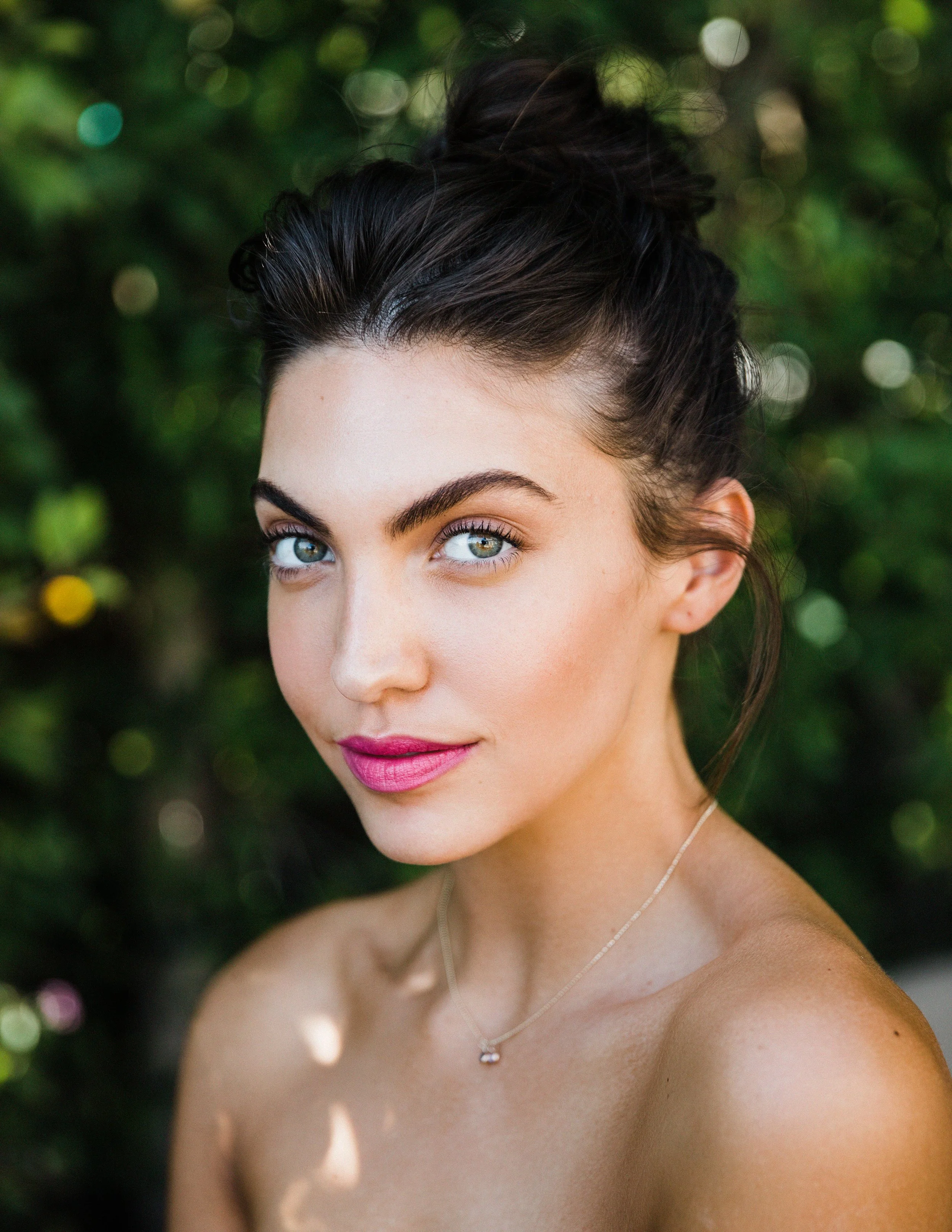 Close-up portrait of a young woman with blue eyes and dark hair styled in an updo, outdoors with a blurred green foliage background.