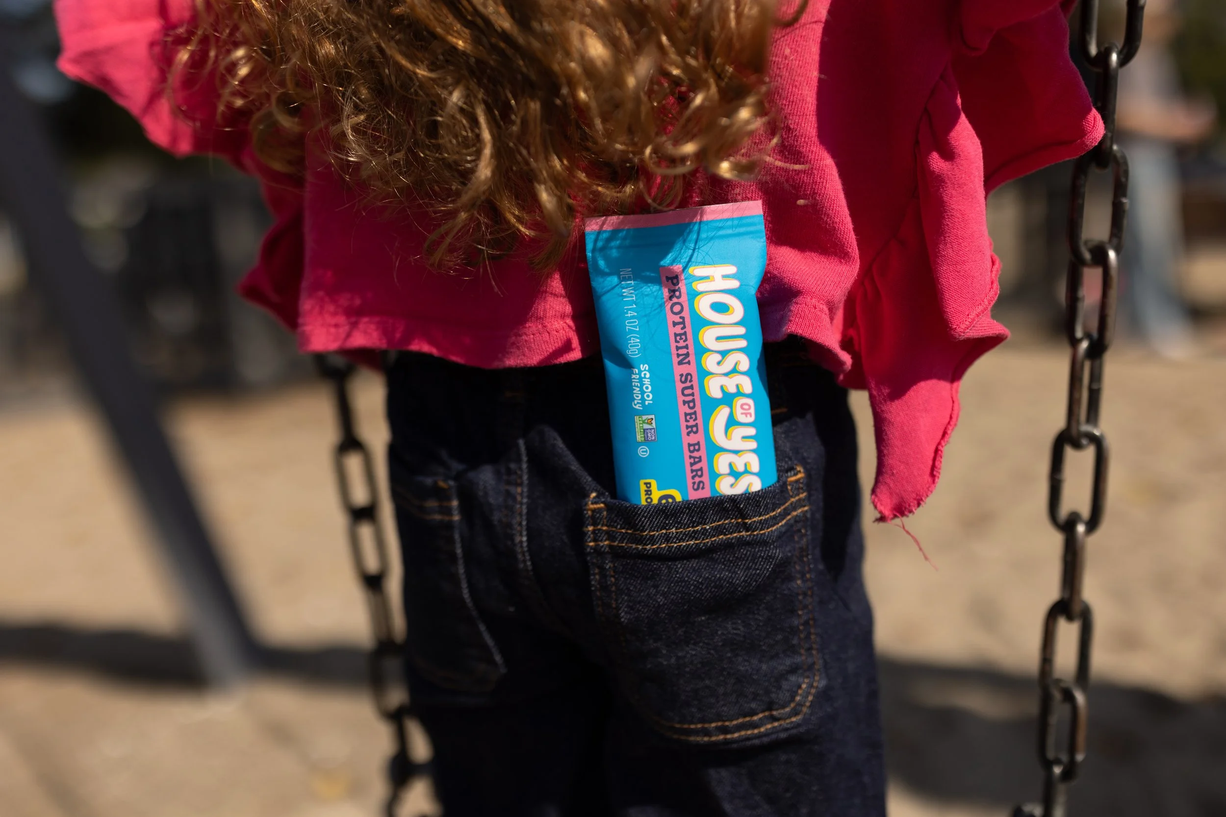 Close-up of a child on a swing, wearing a pink hoodie with a sports team sticker on the chest, and blue jeans.