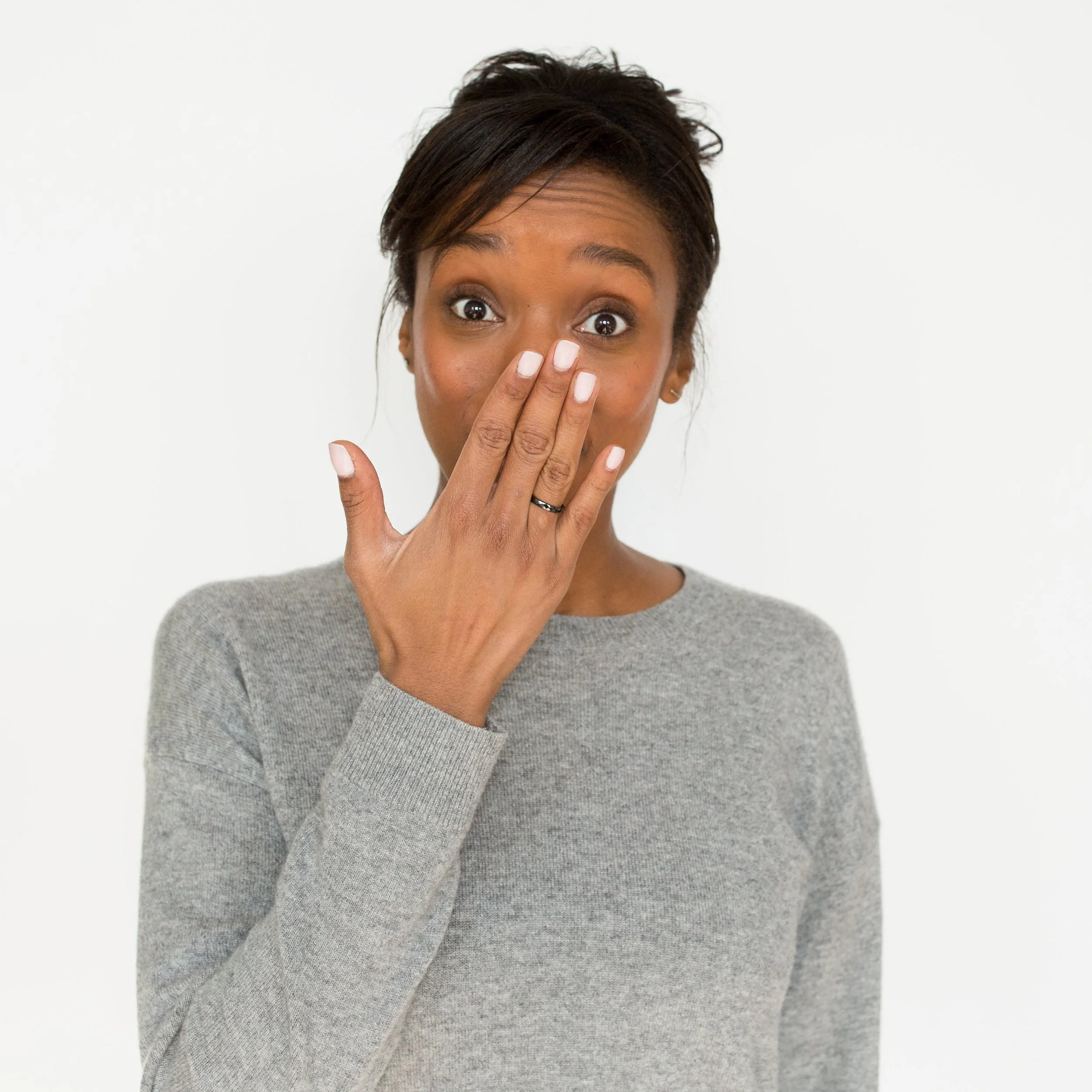 A woman with short dark hair, wearing a gray sweater, covering her nose with her hand, against a plain white background.