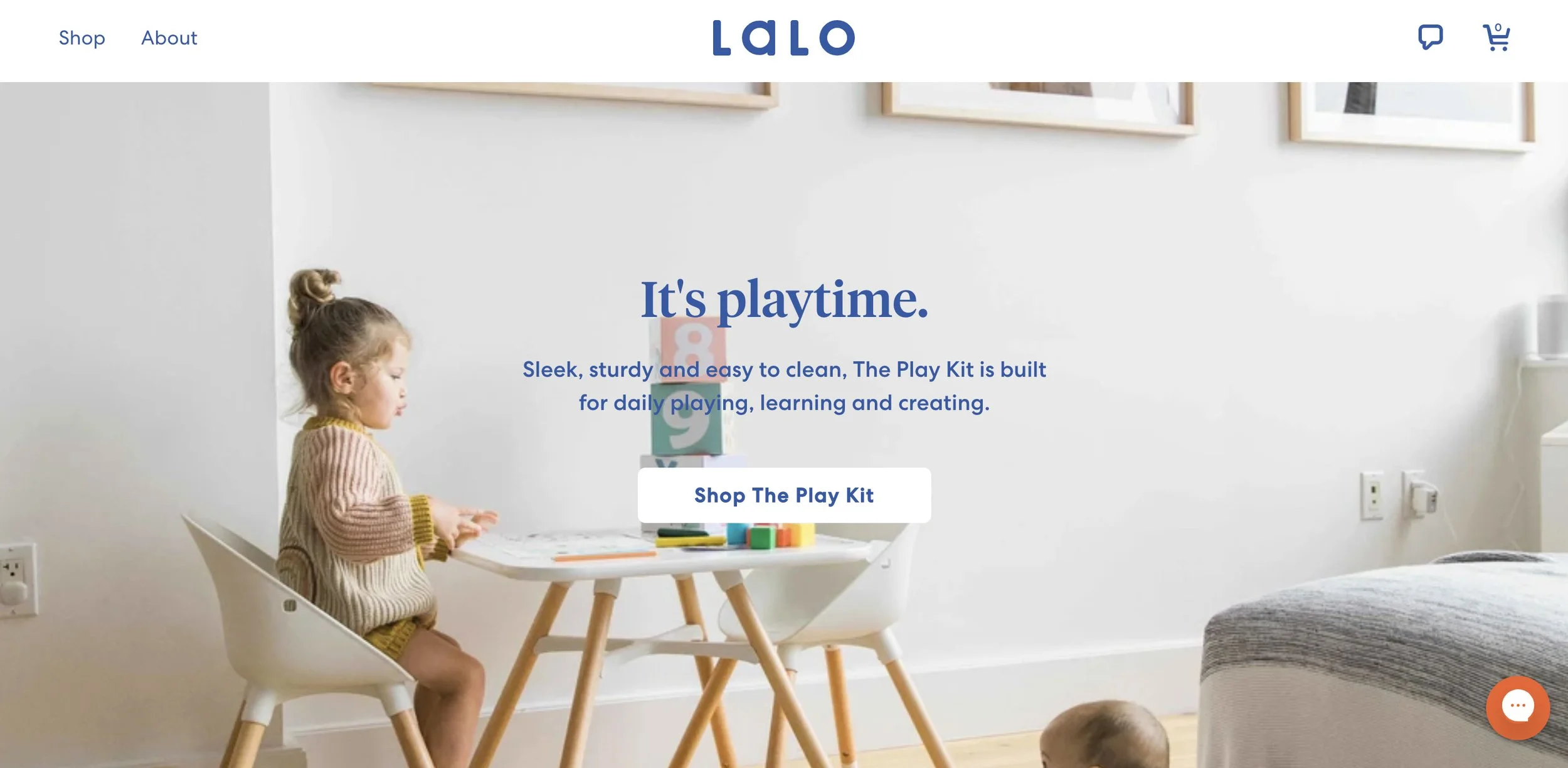 A young girl sitting at a white table with wooden legs, playing with colorful building blocks in a minimalistic room with white walls, framed artwork, and a chair. The scene appears bright and clean, emphasizing play and learning.