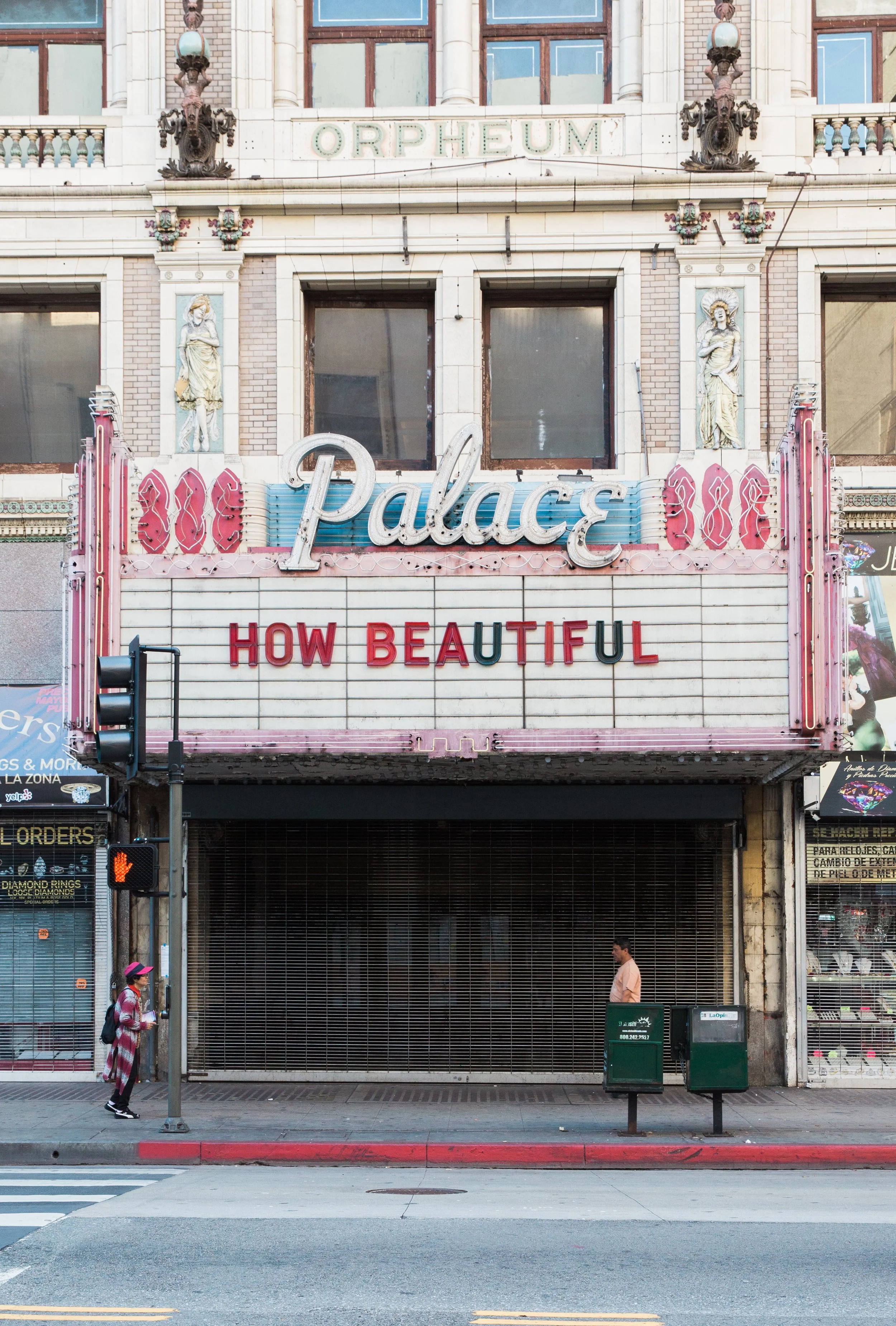 The facade of a building with a sign reading 'Palace' and a marquee with the phrase 'How Beautiful'. There are two statues of women on either side of a window, and the area below the sign is covered with a metal grate. Pedestrians stand and walk acro