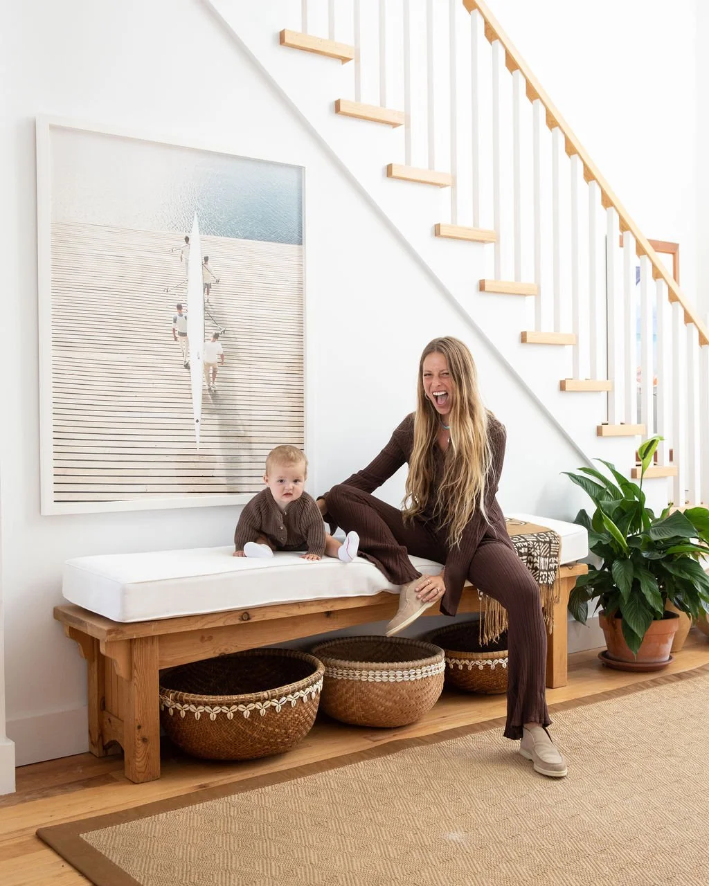 A woman and a young child sitting on a wooden bench inside a modern home, laughing and playing. There is a large plant and a staircase in the background.
