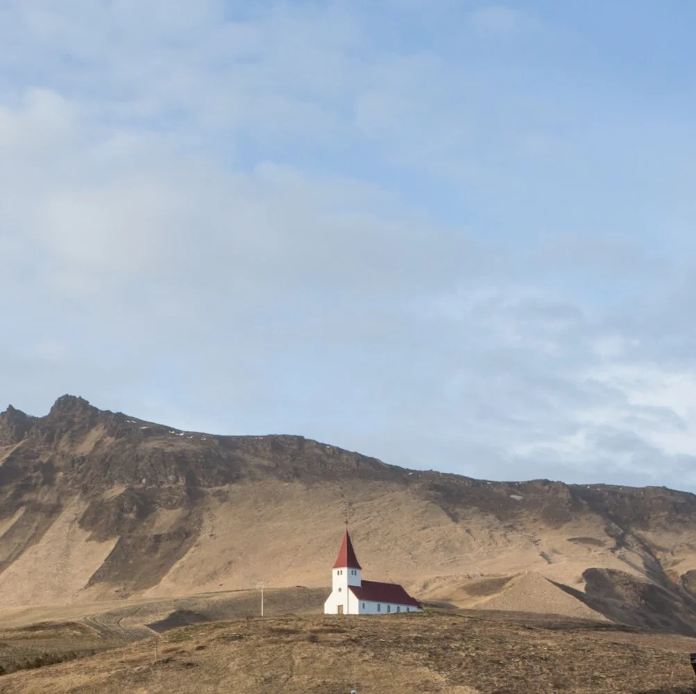 A small white church with a red roof and steeple situated on a grassy hill in front of a mountain range under a partly cloudy sky.