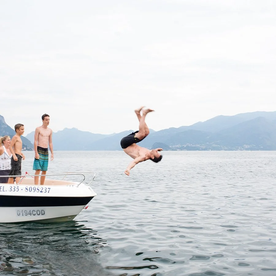 A man jumping off a boat into a lake with three children watching. Mountains are visible in the background, and the sky is cloudy.