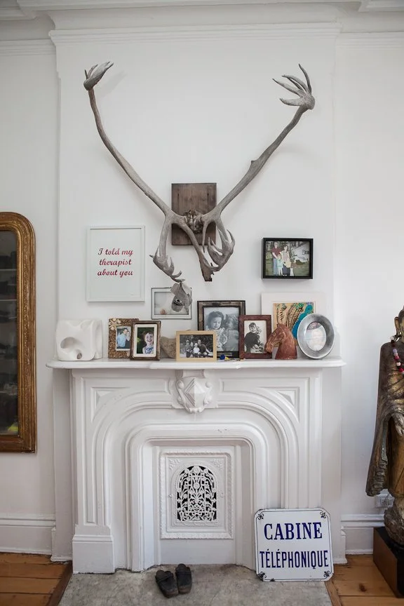 Decorative white fireplace mantle with several framed photographs, a white skull sculpture, a carved wooden horse head, a circular mirror, and a small dog figure on top. Above the mantle hangs a large deer antler set mounted on a wooden plaque. To th