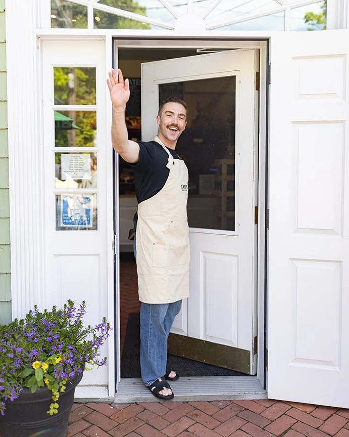 A man smiling and waving from the door of a small shop, wearing a beige apron and casual clothes, outside a white building with a flower pot nearby.