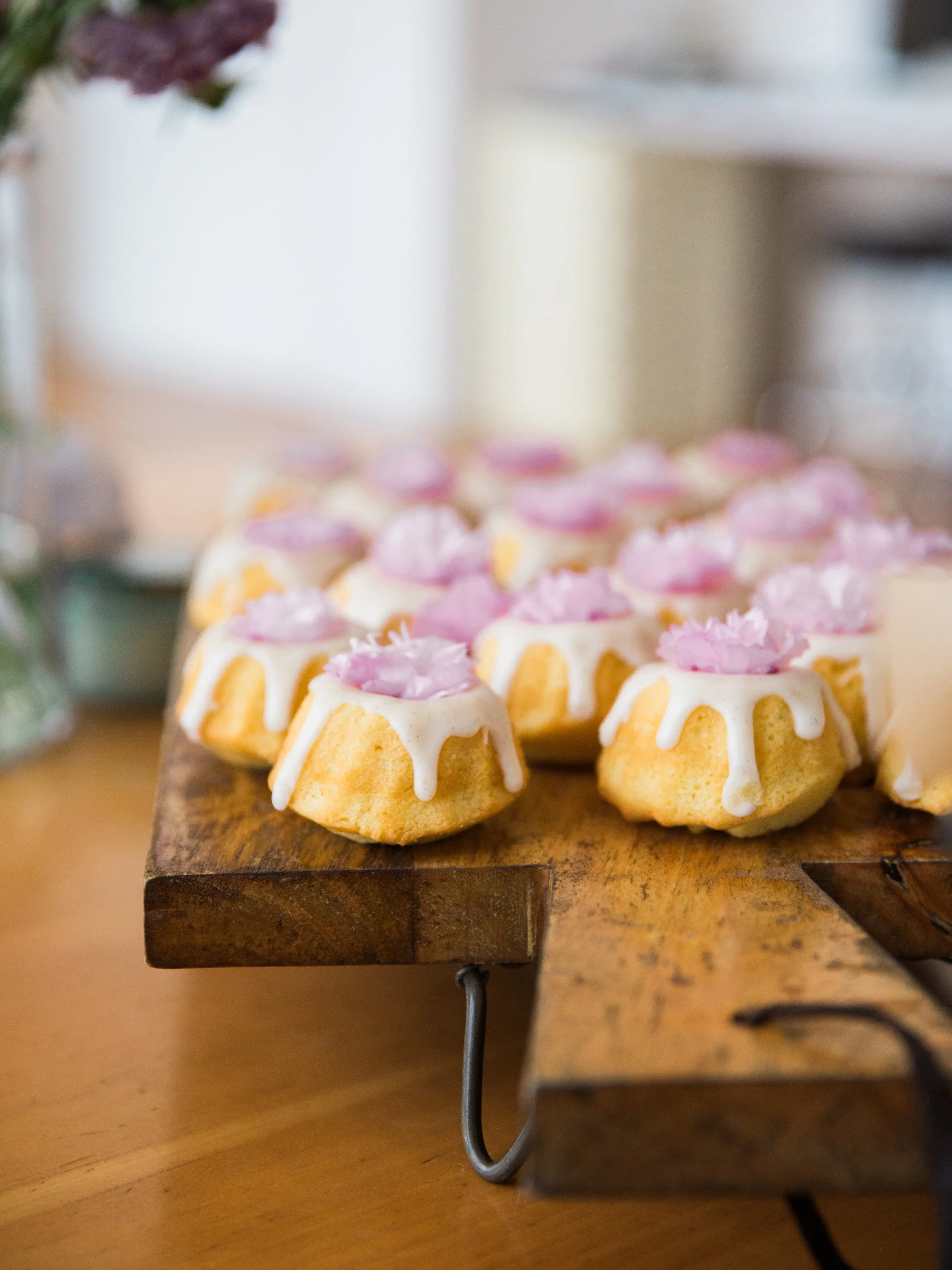 A rustic wooden board with small decorated cupcakes with white icing and pink flowers on top, placed on a wooden table.