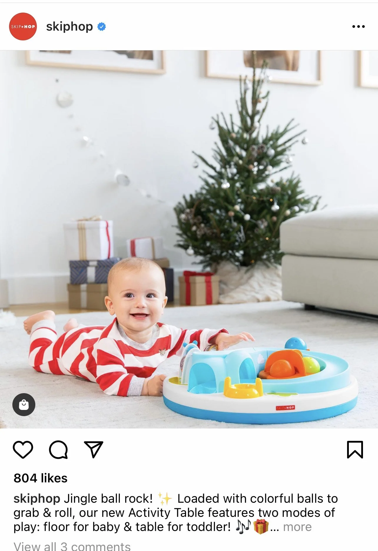 A smiling baby lying on the floor in front of a colorful activity table with toy balls, with a decorated Christmas tree and wrapped presents in the background.