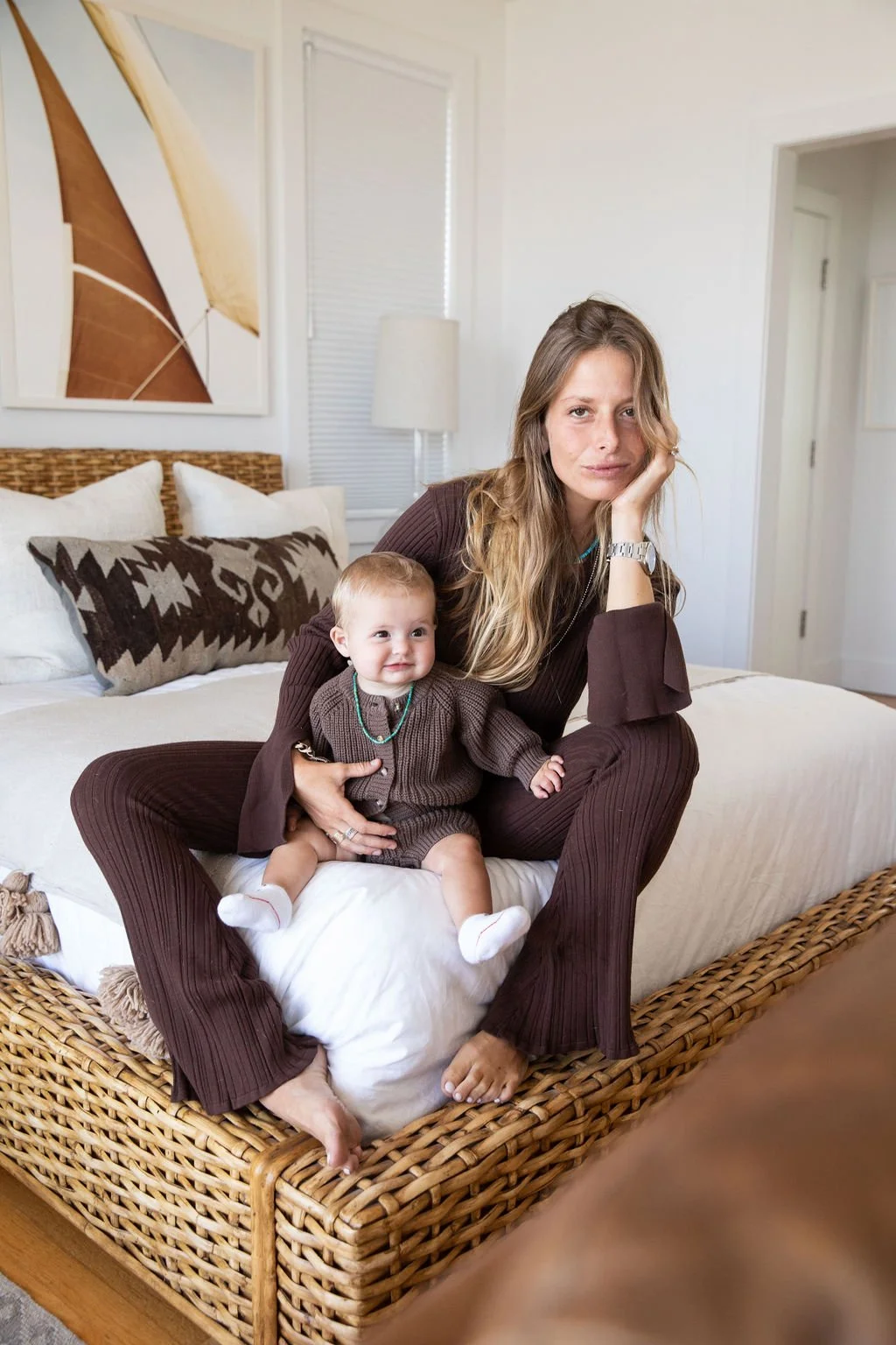 A woman sitting on a wicker bed with a young baby on her lap, both dressed in matching brown outfits, in a cozy room with white walls and artwork.