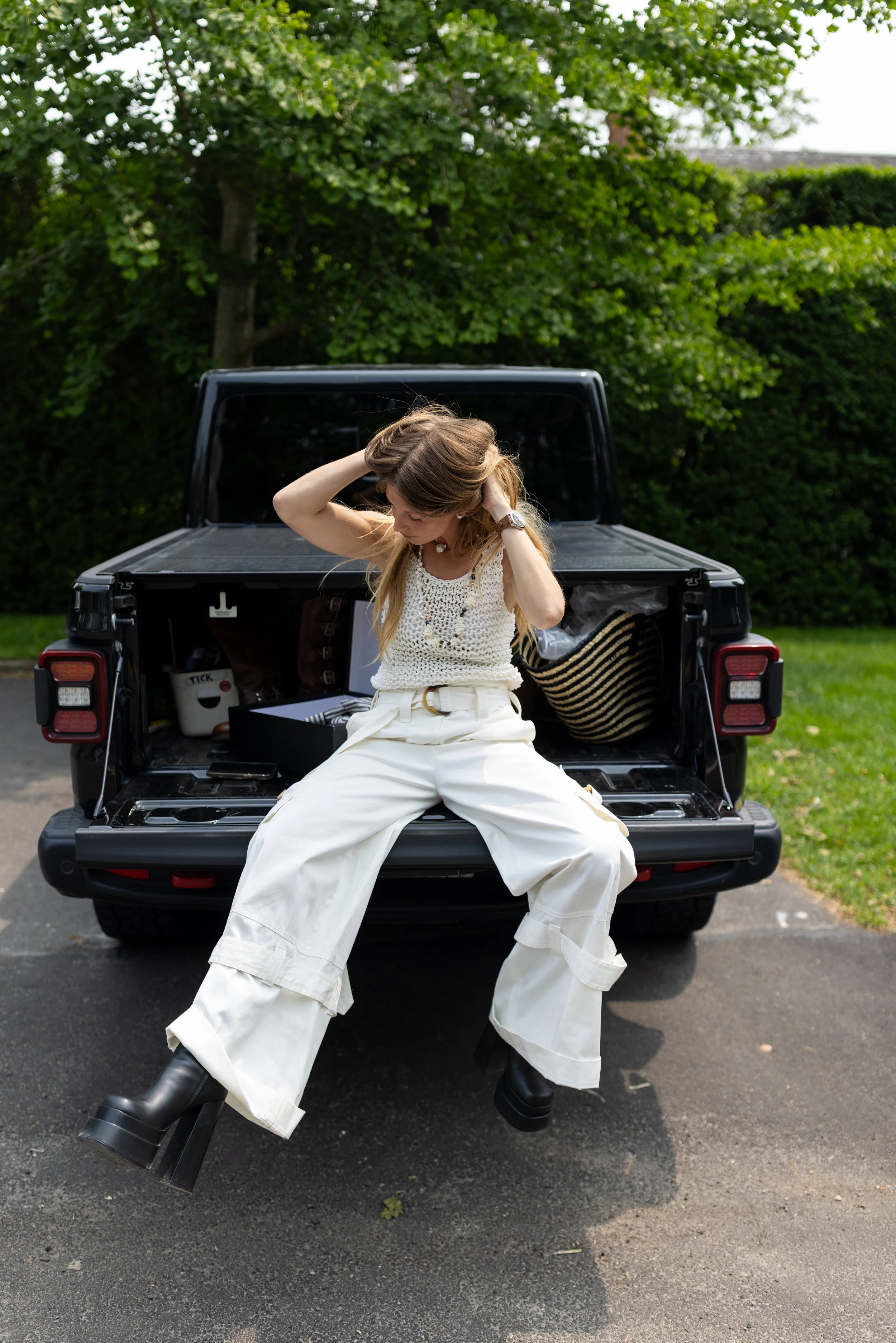 A woman with long blonde hair, wearing a white crochet top, high-waisted white pants, and black boots, sitting on the open tailgate of a black pickup truck. She is adjusting her hair and looking down, with a strappy bracelet on her left wrist. The tr