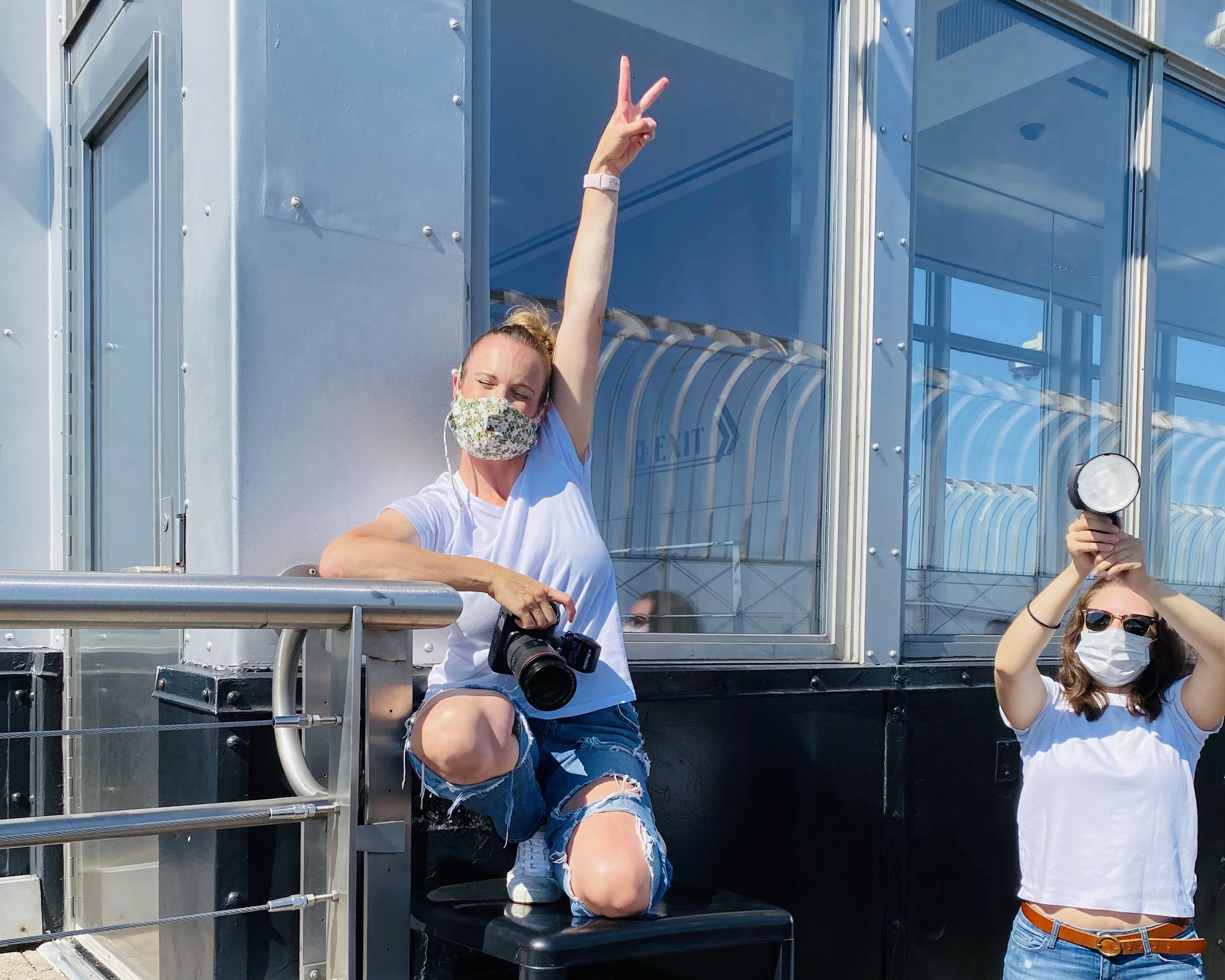 A woman wearing a floral face mask, white t-shirt, and ripped jeans sitting on a black stair, making a peace sign with her hand. A young girl wearing a white t-shirt, sunglasses, and a white face mask is also raising her arm, holding a flashlight. They are outdoors in front of a shiny, metallic building with glass windows.