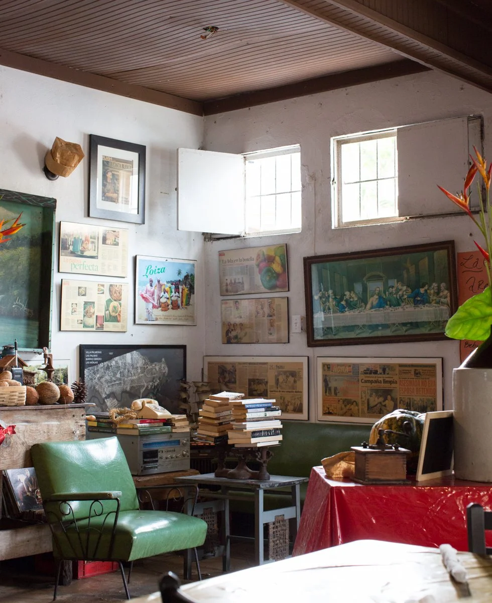Eclectic interior room with white walls, wooden ceiling, and two small windows; decorated with framed vintage posters, newspapers, and a large painting of The Last Supper; contains a green armchair, stacks of books, a red tablecloth, and various deco