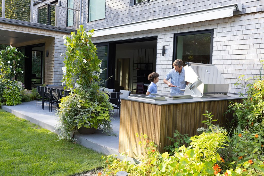 Man and boy at an outdoor kitchen with a built-in grill, on a patio with plants and outdoor furniture.