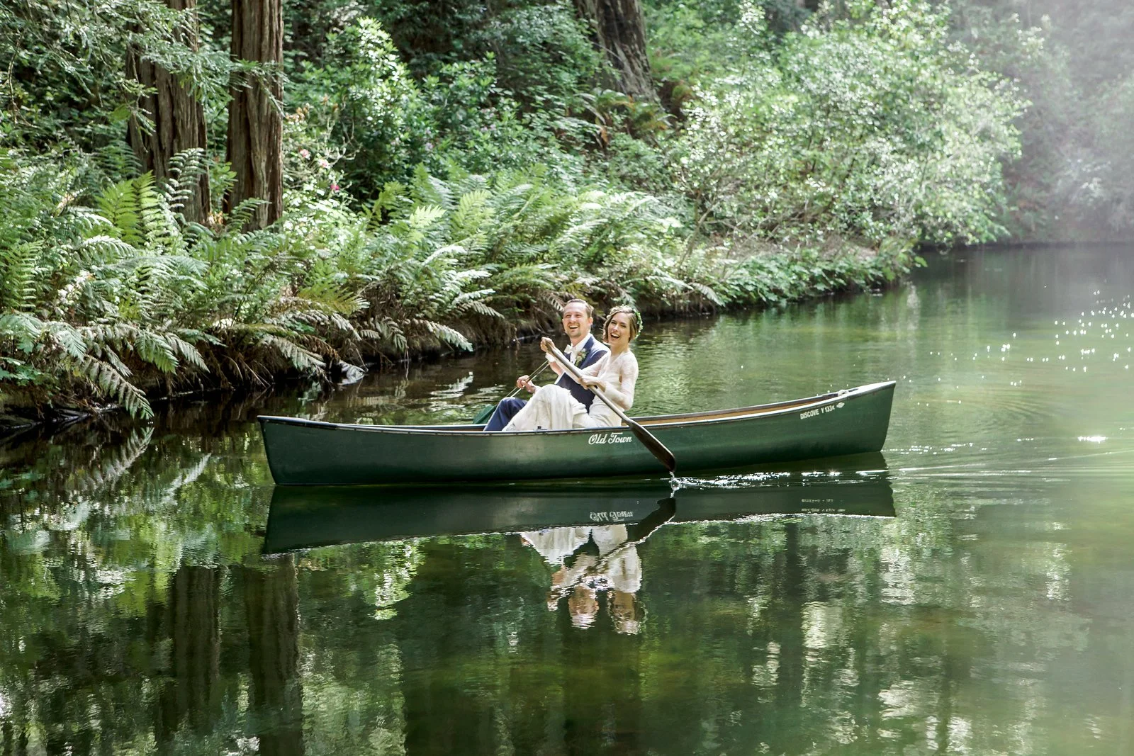 A happy couple dressed in wedding attire rowing a green canoe on a calm, forested river.