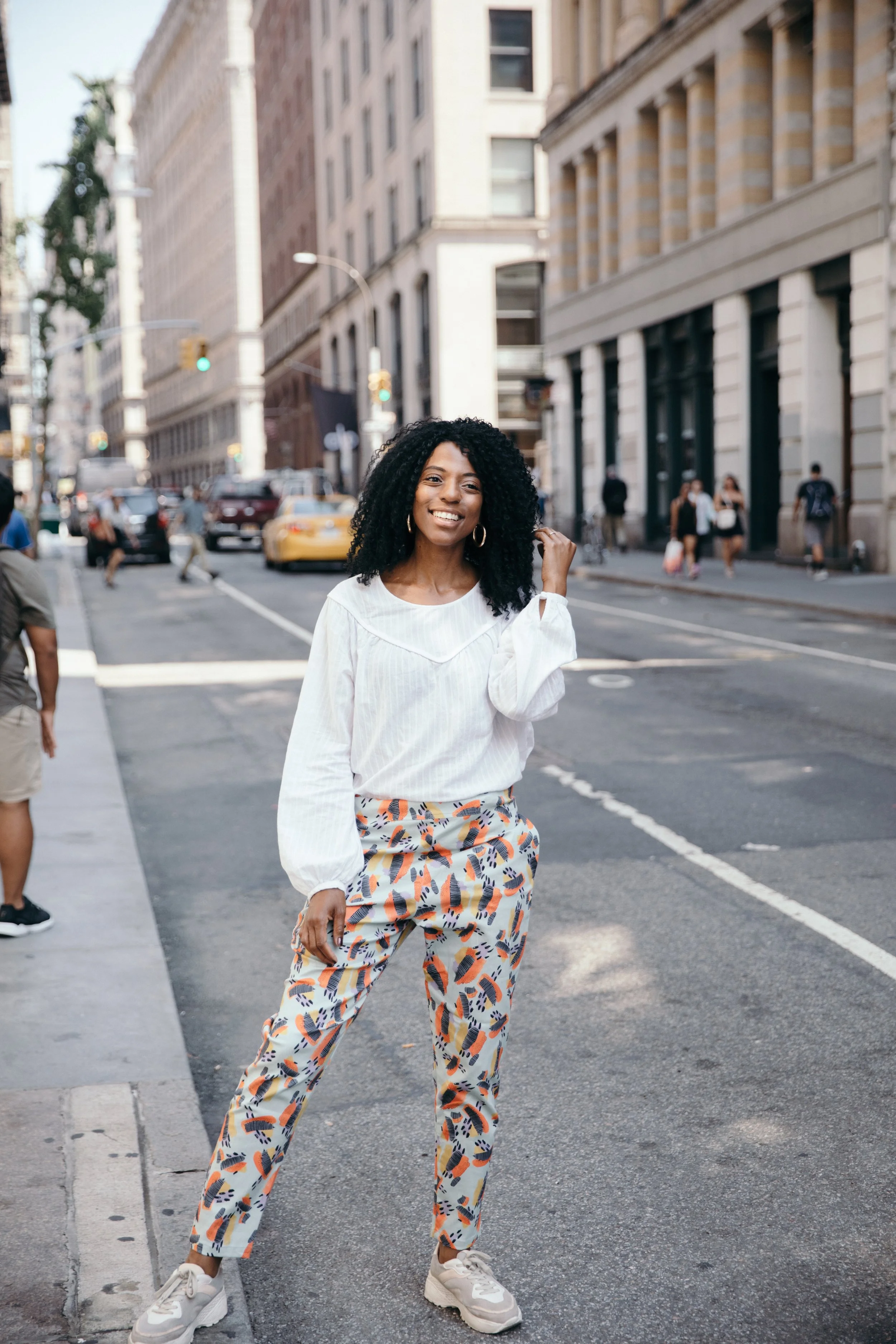 A young woman with curly black hair, wearing a white blouse and colorful patterned pants, smiling and standing in the middle of a city street with tall buildings, cars, and pedestrians in the background.