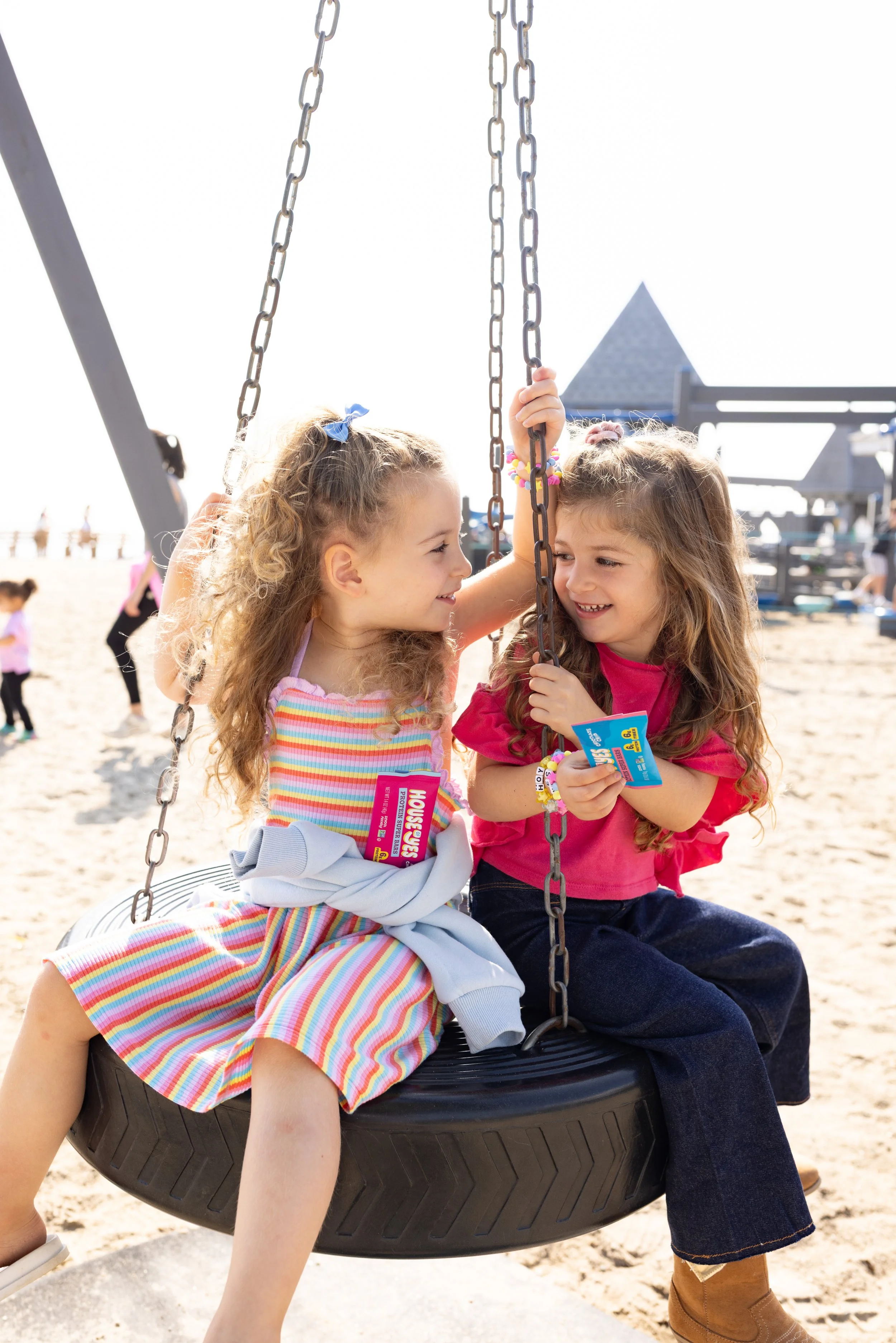 Two young girls with curly hair sitting on a tire swing at the beach, smiling and hugging each other.