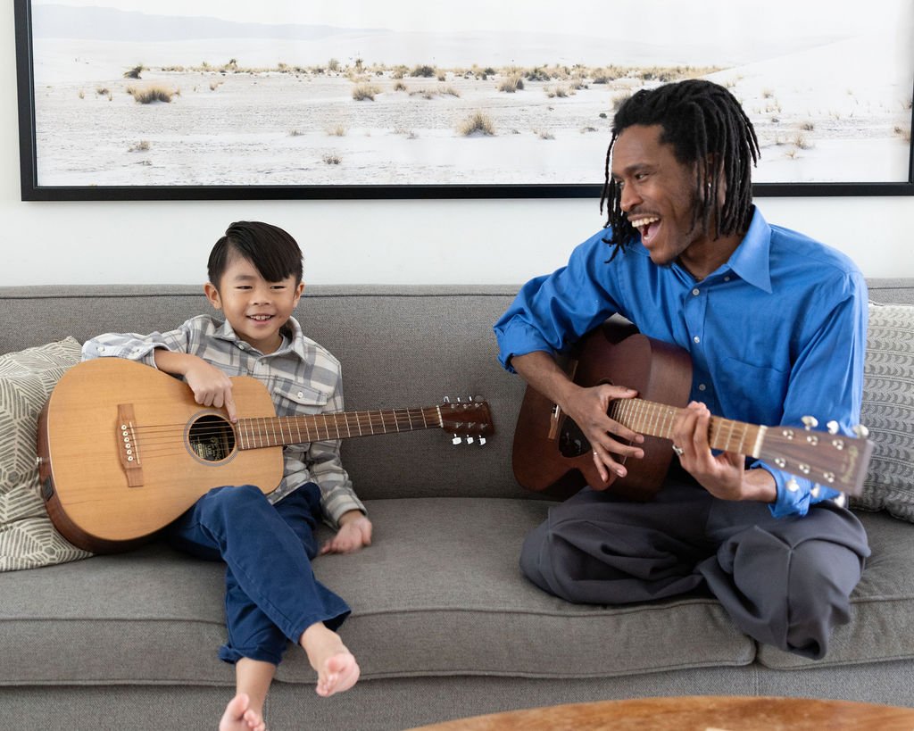 A young boy and an adult man playing guitars and singing together on a gray couch in a living room.