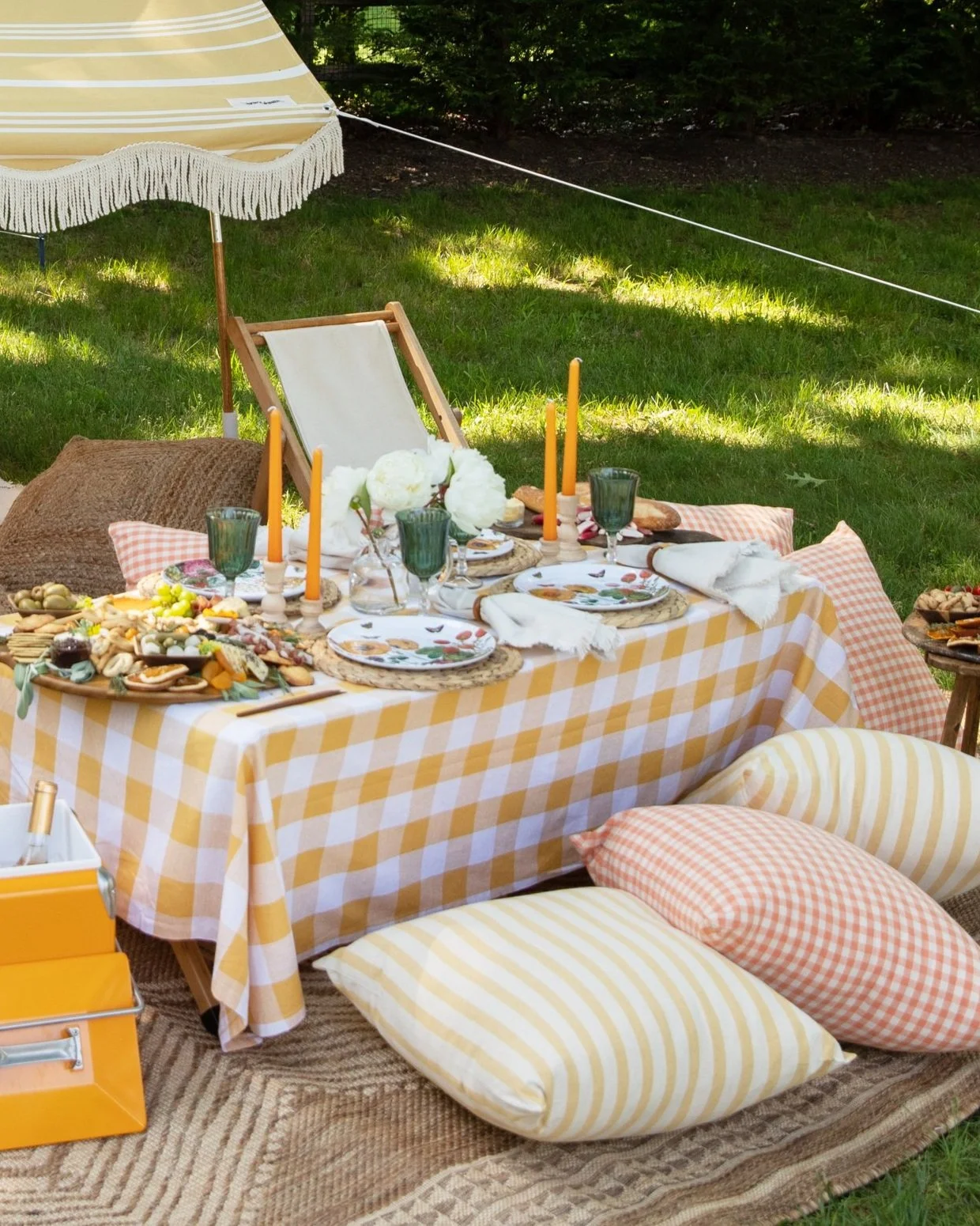 Outdoor picnic table setup with yellow checkered tablecloth, plates, glasses, candles, and snacks, surrounded by cushions and pillows on a grassy area with trees in the background