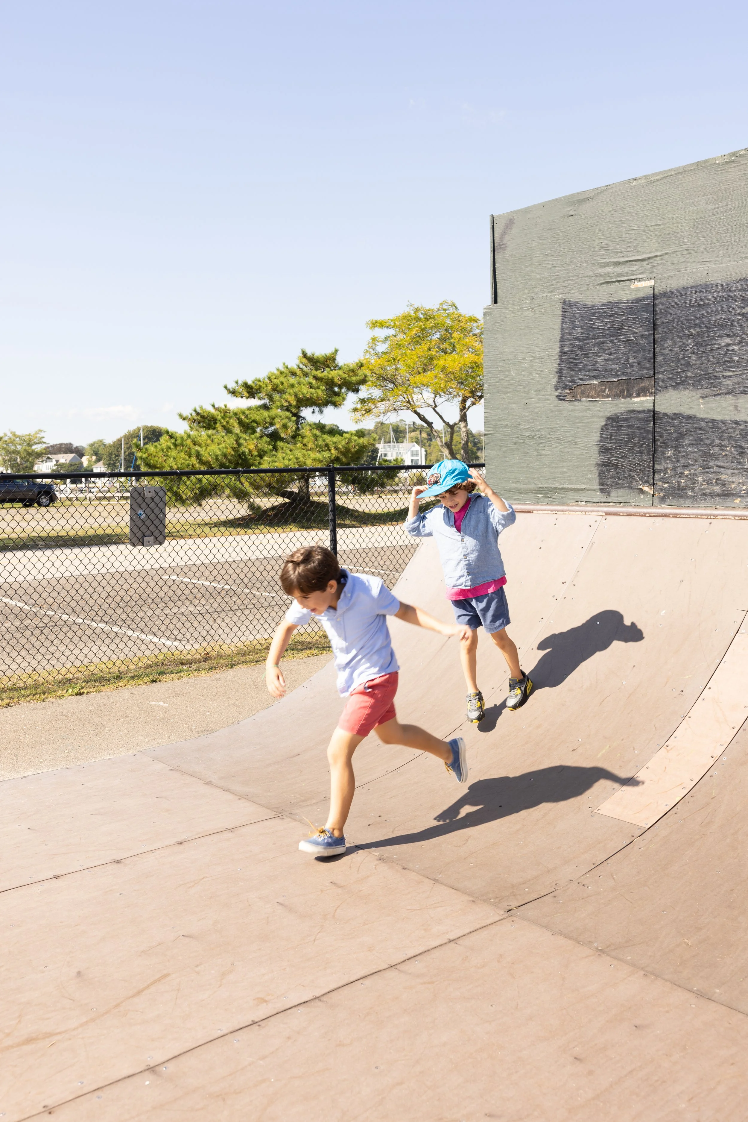 Two children running down a skate ramp at a skatepark, with a fence, trees, and buildings in the background.