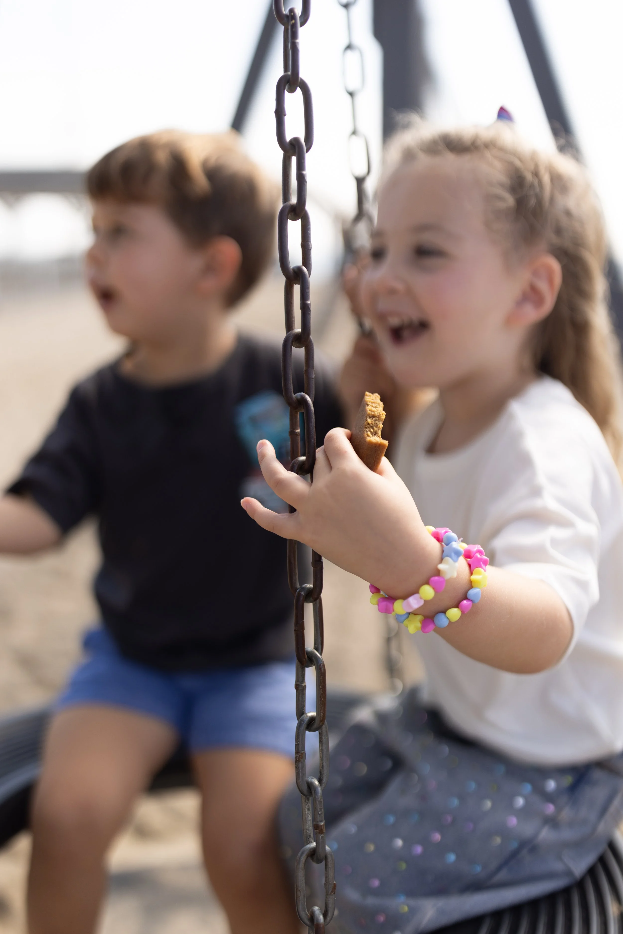 Two young children, a boy and a girl, sitting on a swing set outdoors. The girl is holding a cookie and wears colorful beaded bracelets. They are both smiling and seem to be enjoying their time.