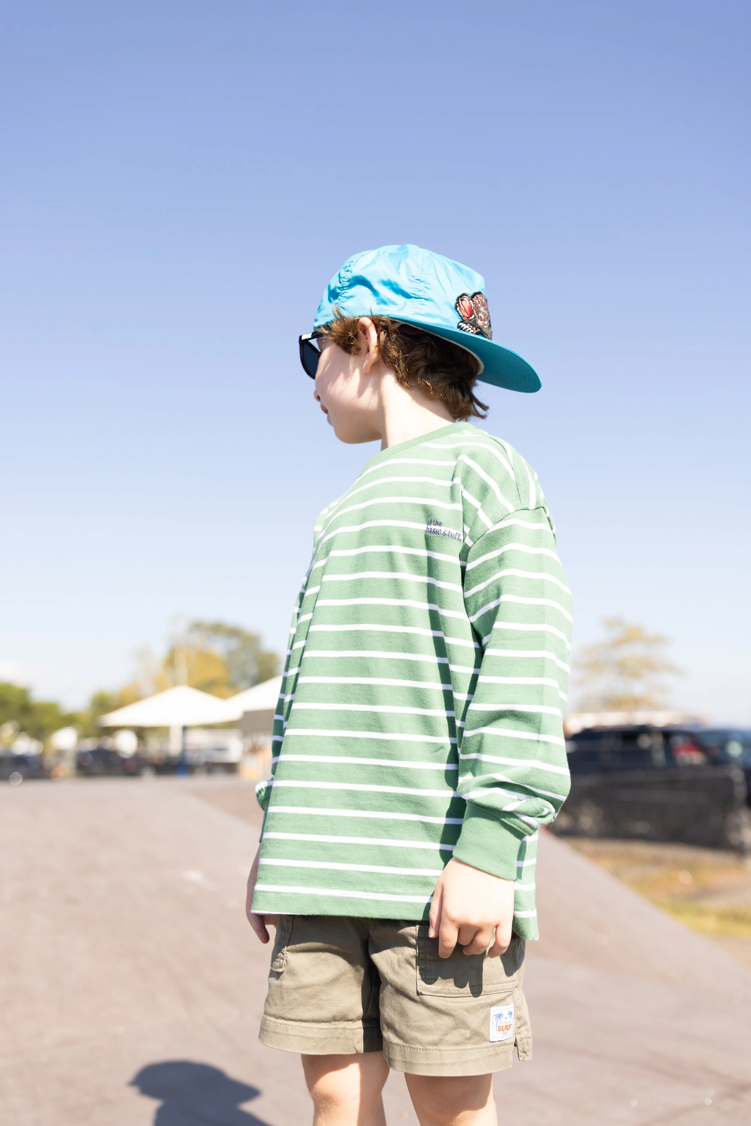 A person wearing a green and white striped t-shirt, khaki shorts, a blue cap with a patch, and sunglasses, standing outdoors on a sunny day with a parking lot and tents in the background.