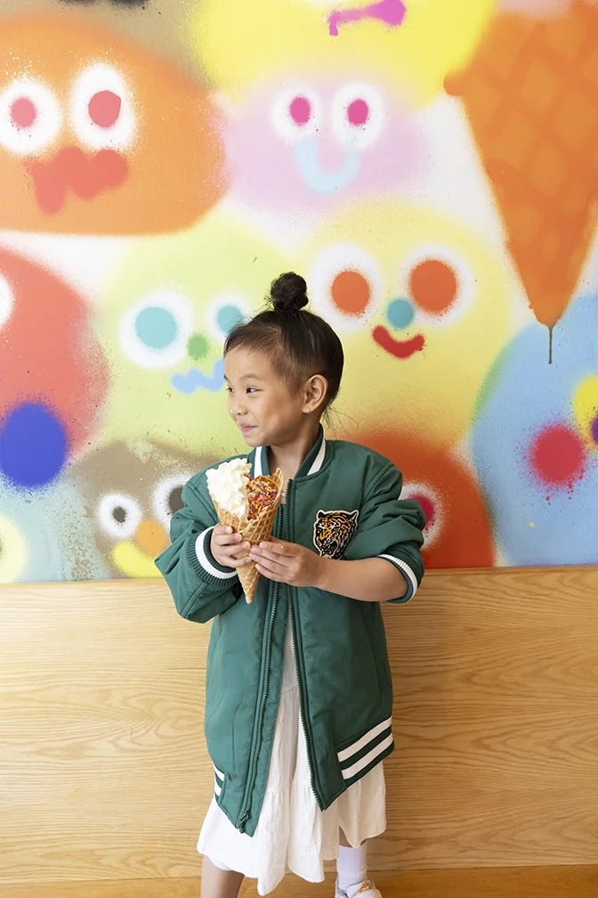 A young girl with a top knot hairstyle wearing a green jacket with a tiger patch, holding an ice cream cone, standing against a colorful graffiti wall with cartoon faces.
