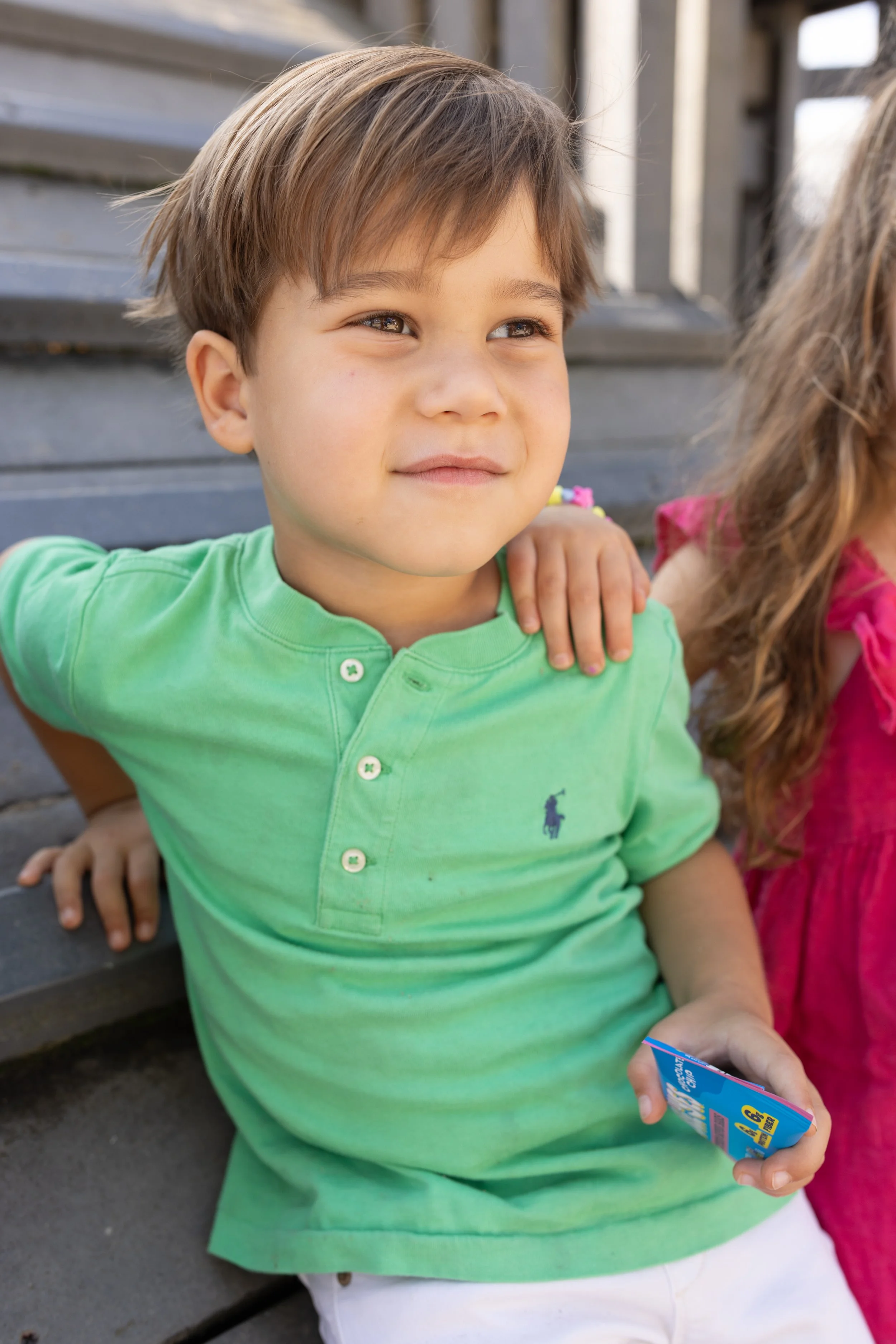 Close-up of a young boy with brown hair, wearing a green polo shirt, sitting outdoors near stairs, holding a blue item, with another girl partially visible beside him.