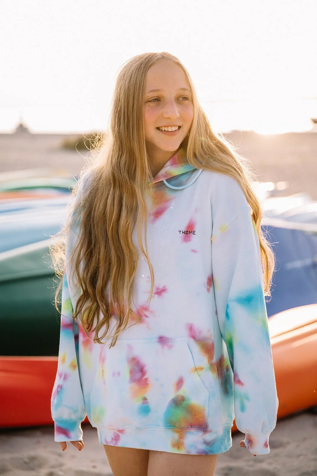 A young woman with long wavy blonde hair stands outdoors in front of colorful kayaks, wearing a tie-dye hoodie with the word 'THEME' on it, smiling as the sun sets.