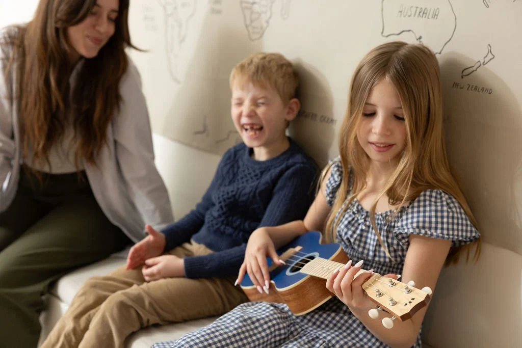 A young girl with red hair playing a small acoustic guitar while sitting on a bench between a boy with red hair and an adult woman, with a world map on the wall behind them.