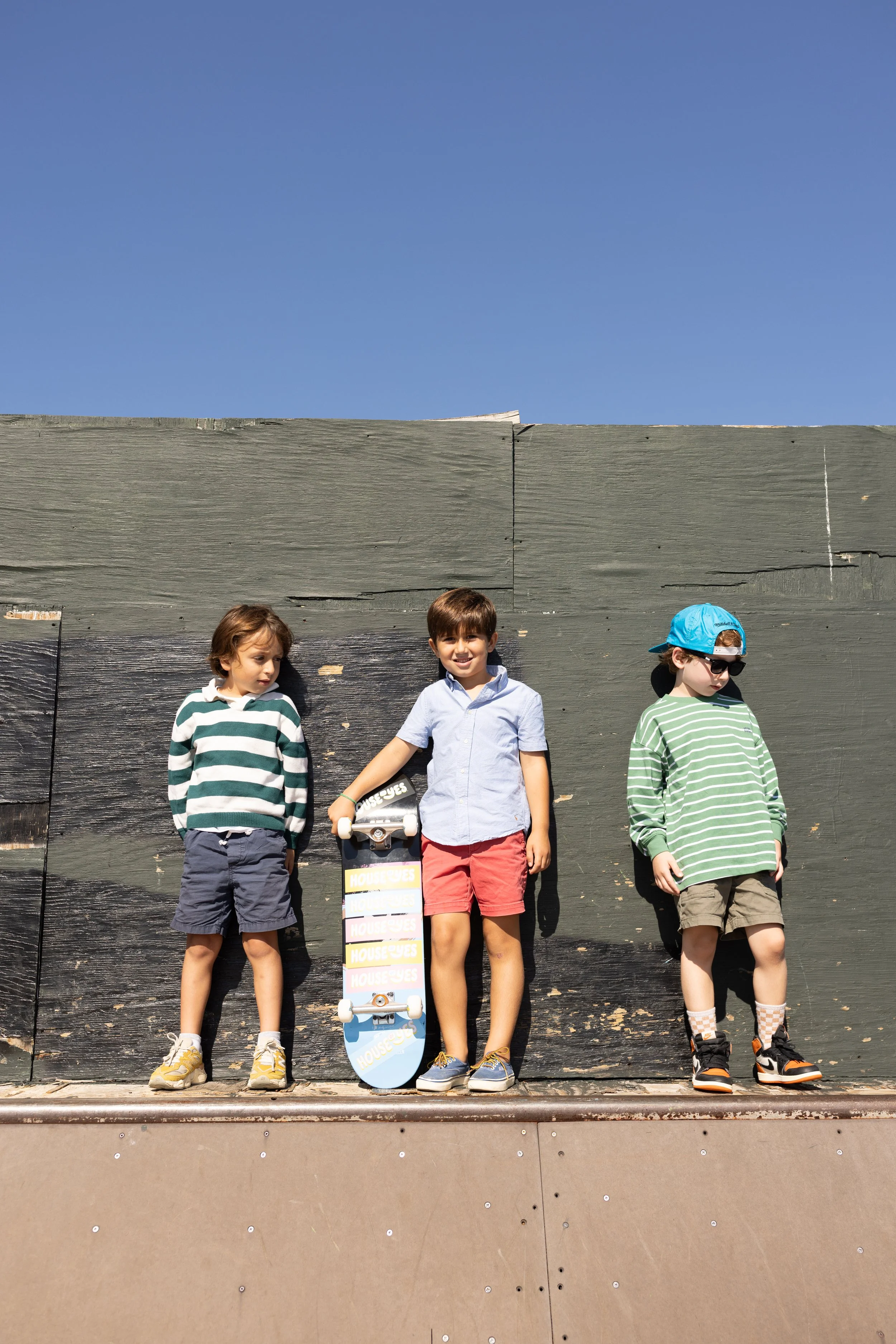 Three boys standing on a skateboard in front of a dark wooden wall outdoors under a blue sky. One boy is holding a skateboard with the word 'House' on it. The boys are dressed in casual summer clothes and sneakers.