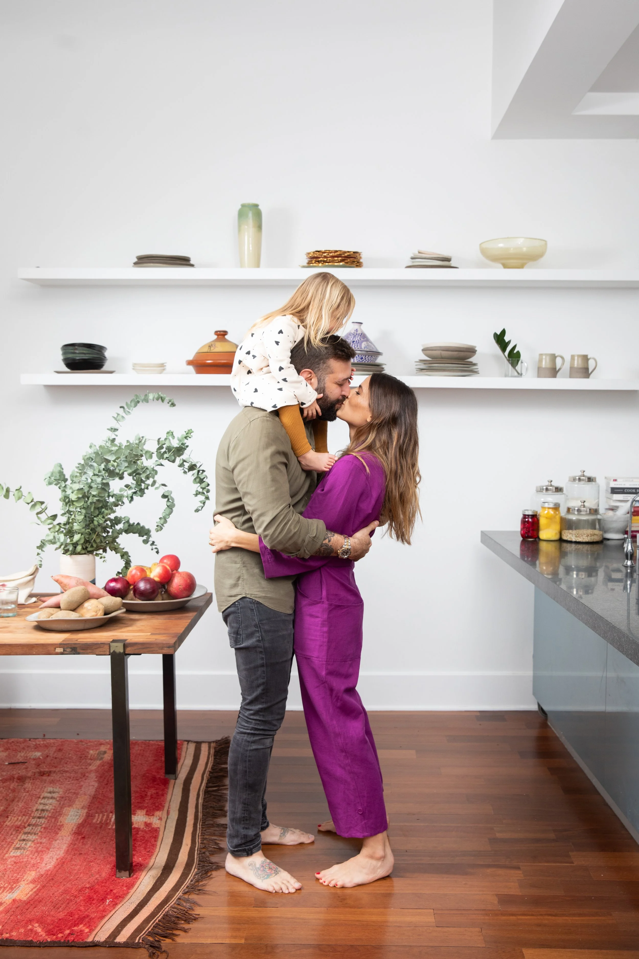 A couple in a kitchen with a child on the husband's shoulders, kissing. The woman and man are barefoot on a hardwood floor, the woman in a purple dress and the man in dark jeans and an olive green shirt. The kitchen has a table with fruits and vegeta