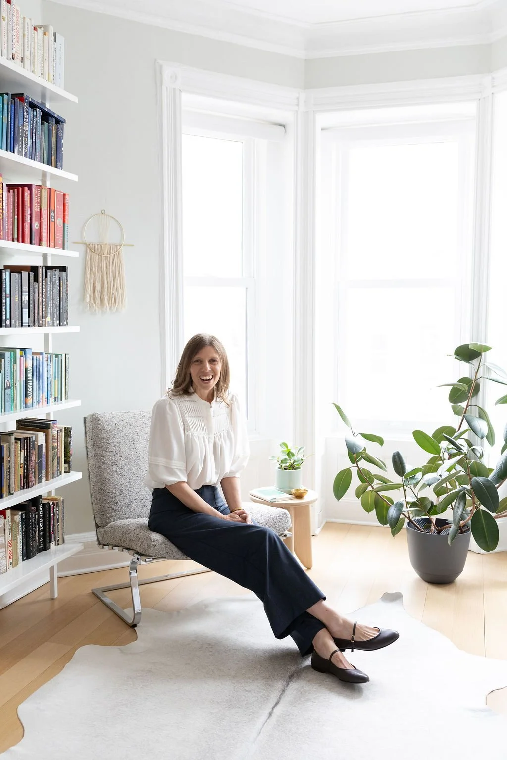 A woman sitting on a gray chair in a bright, white room with large windows, a bookshelf with colorful books on the left, and a large green potted plant on the right. She is smiling and wearing a white blouse and dark pants.