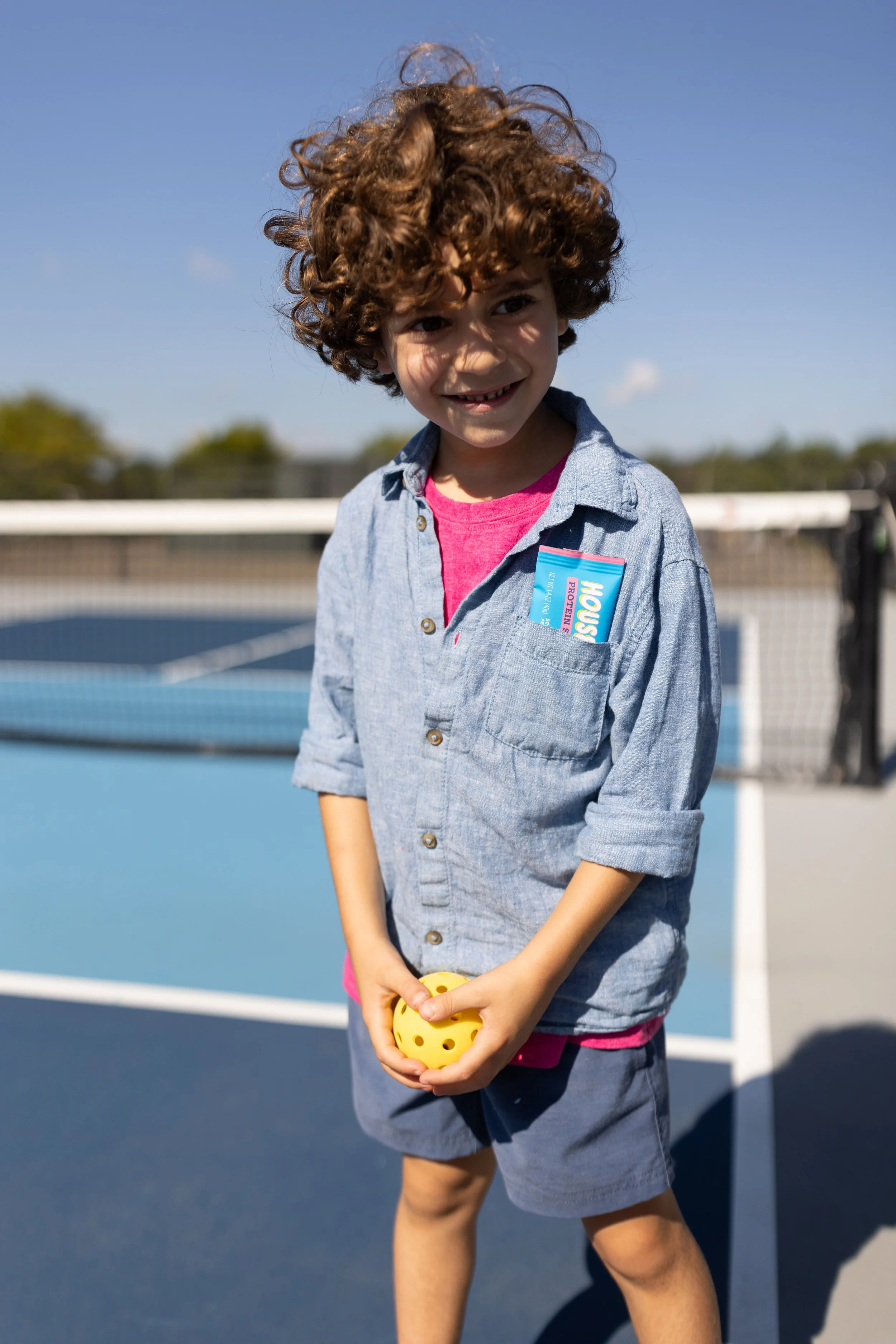 A young boy with curly brown hair smiling outdoors on a tennis court, holding a yellow ball, wearing a blue jacket over a pink shirt.