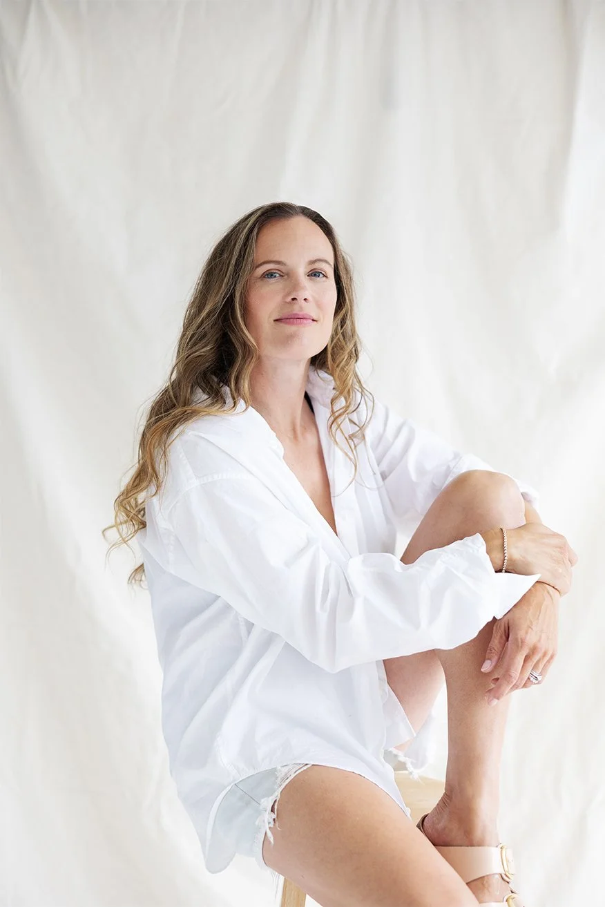 A woman with long wavy hair wearing a white shirt, sitting on a stool, with a light-colored background.