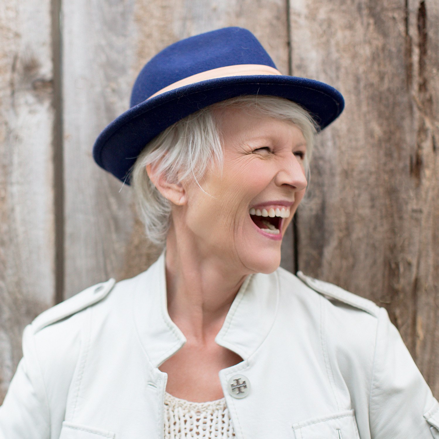 Woman with short gray hair wearing a blue hat and white jacket, smiling and laughing outdoors.