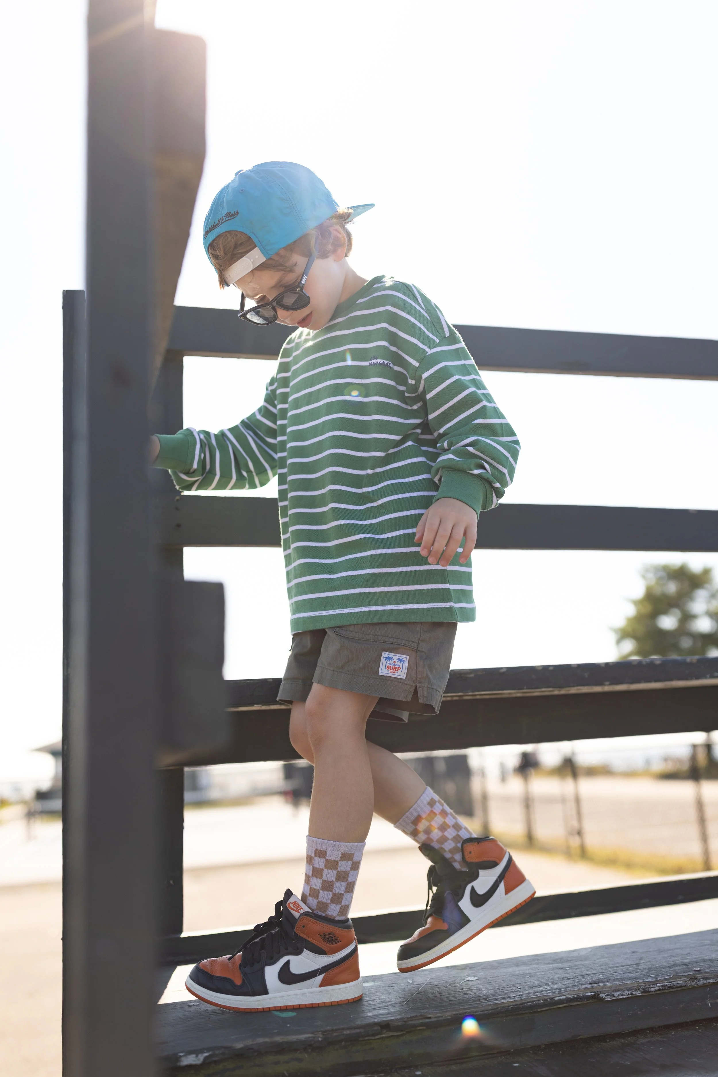 Young boy wearing a blue cap, striped green and white shirt, checkered socks, and sneakers walking down a wooden ramp with black railings at a park.