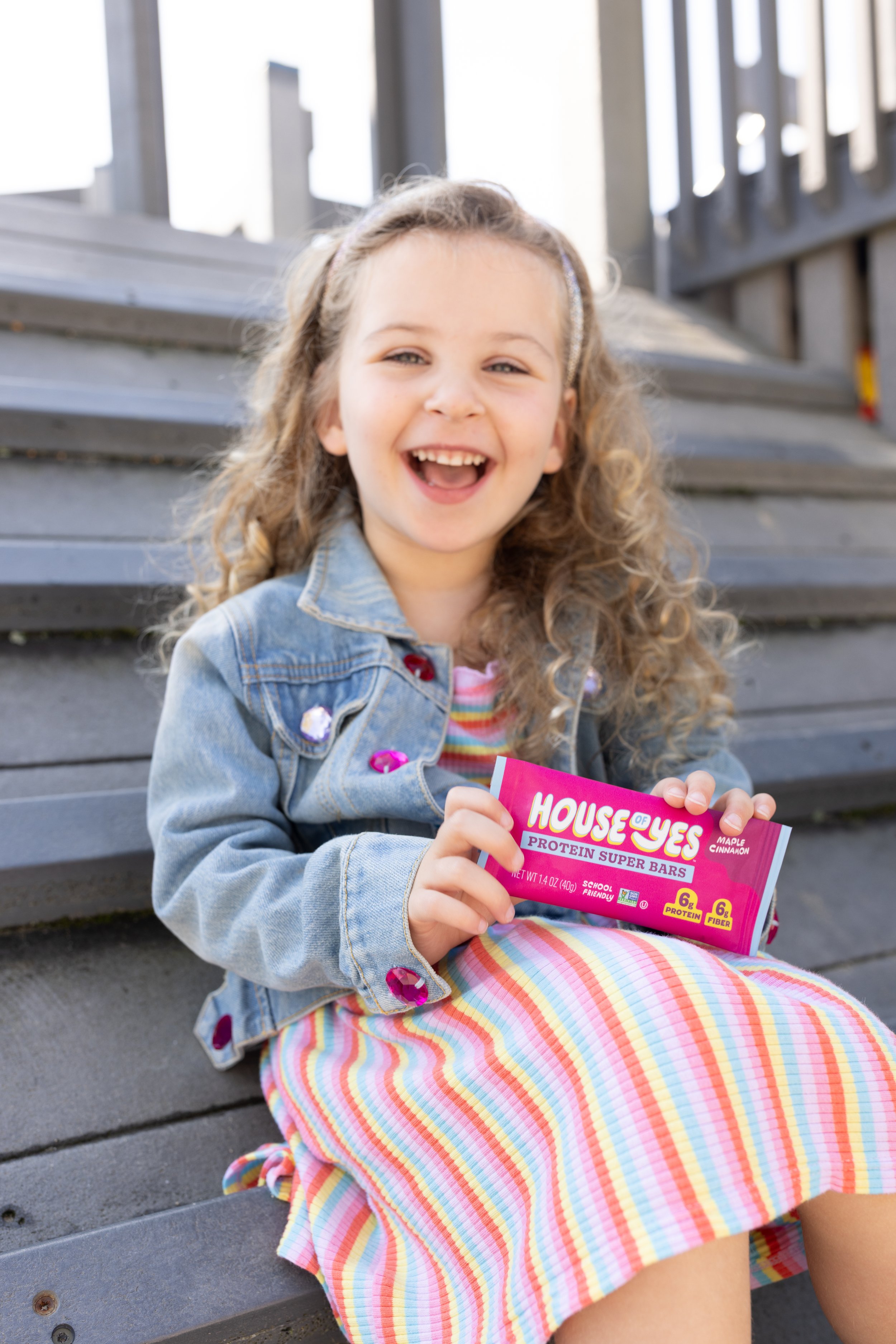 A young girl with curly hair, wearing a denim jacket with pink sequin details, sitting on outdoor steps, smiling and holding a pink packet of House of Yes protein super bars.