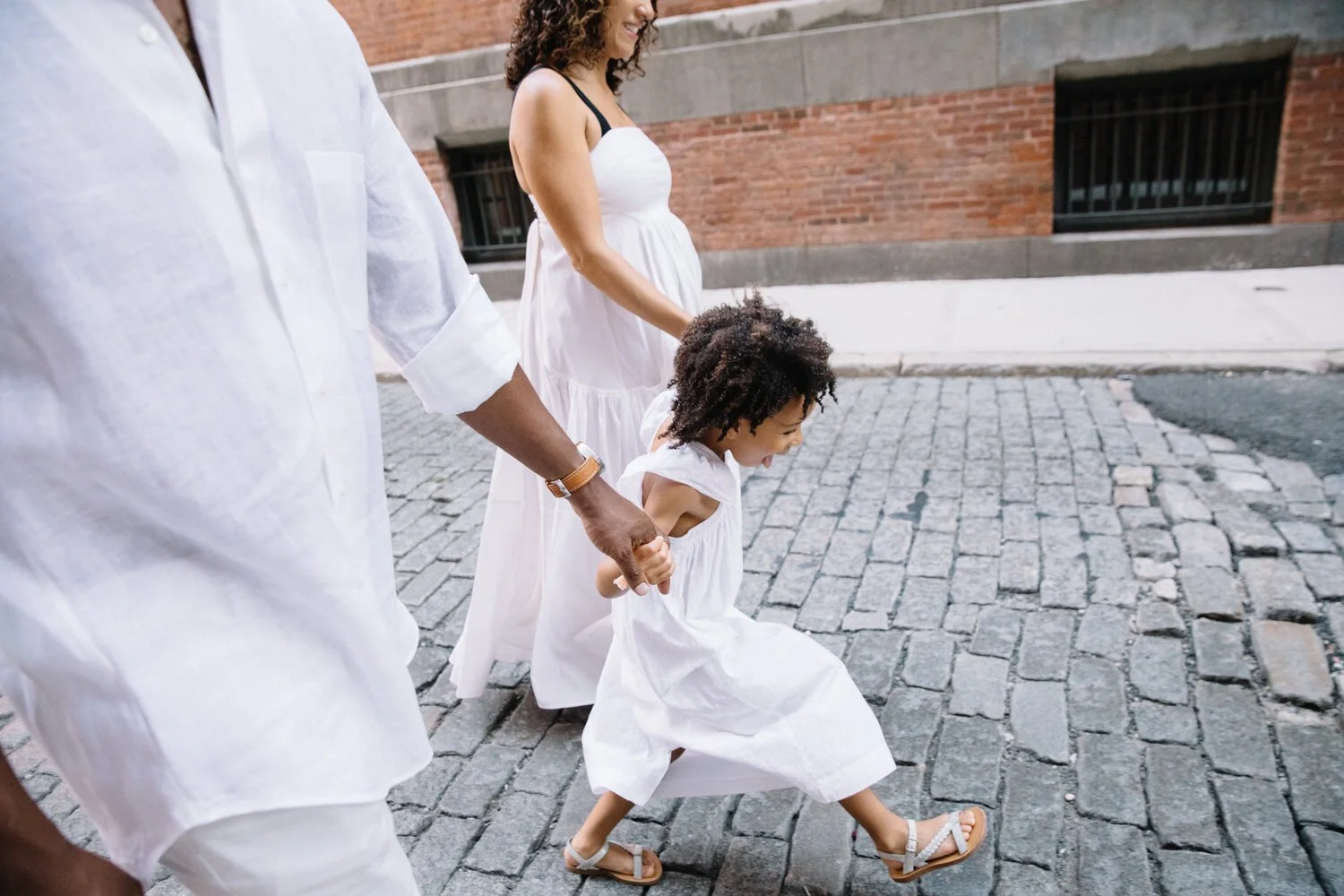 A family walking on a cobblestone street, with a woman and a young girl holding hands, both dressed in white dresses, and a man also dressed in white.