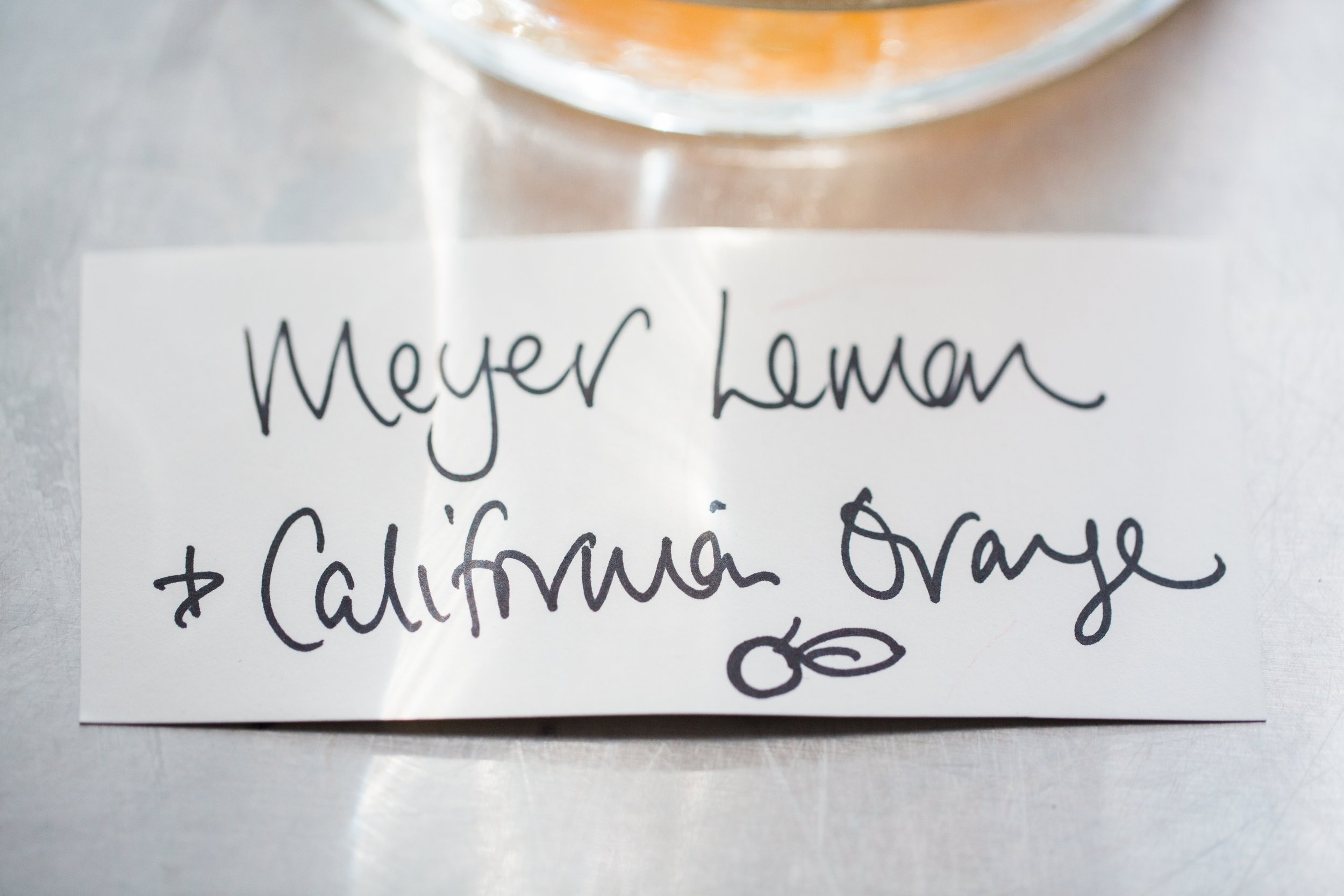 Close-up of a handwritten note that says "Meyer Lemon" followed by additional text, with a glass containing a liquid placed on top of the note.