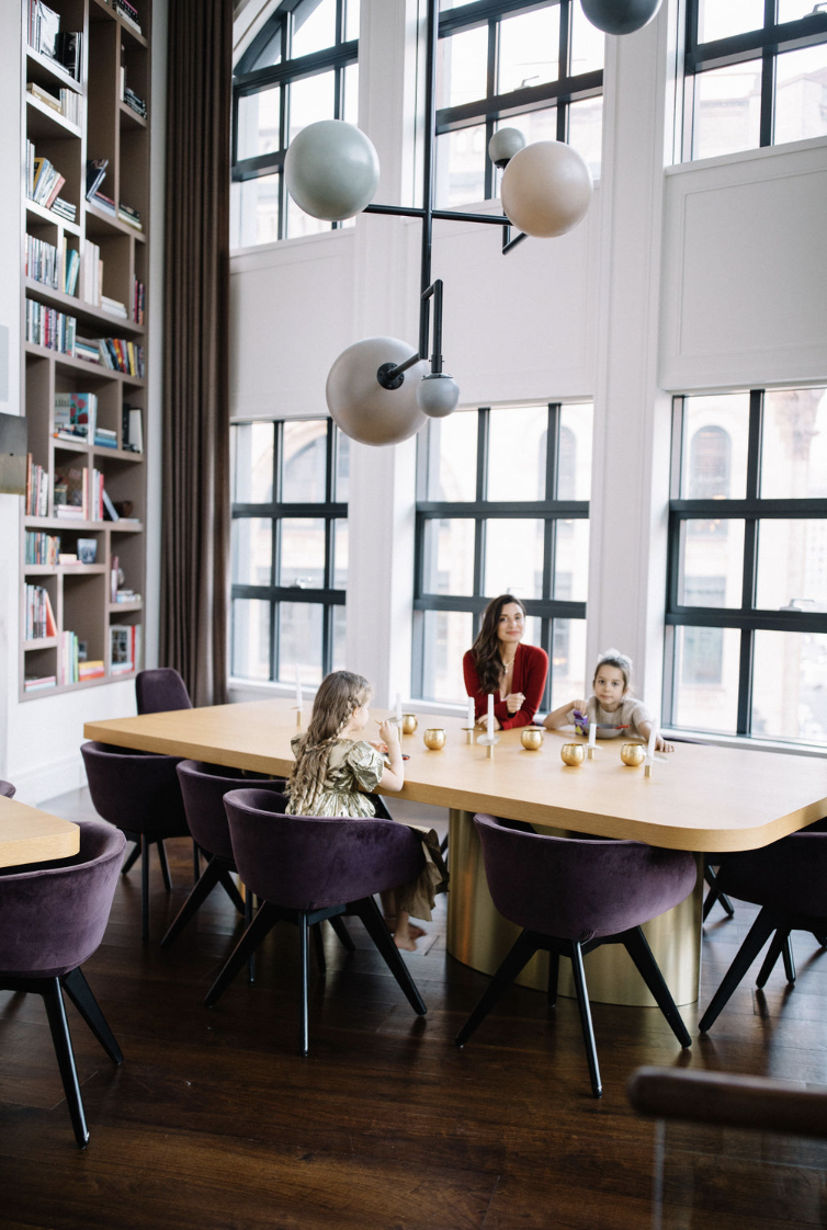 A woman and two young girls sitting at a large wooden dining table in a bright, modern room with large windows. The table has small candles or vases, and the chairs are upholstered in purple. There is a tall bookshelf filled with books on the left si