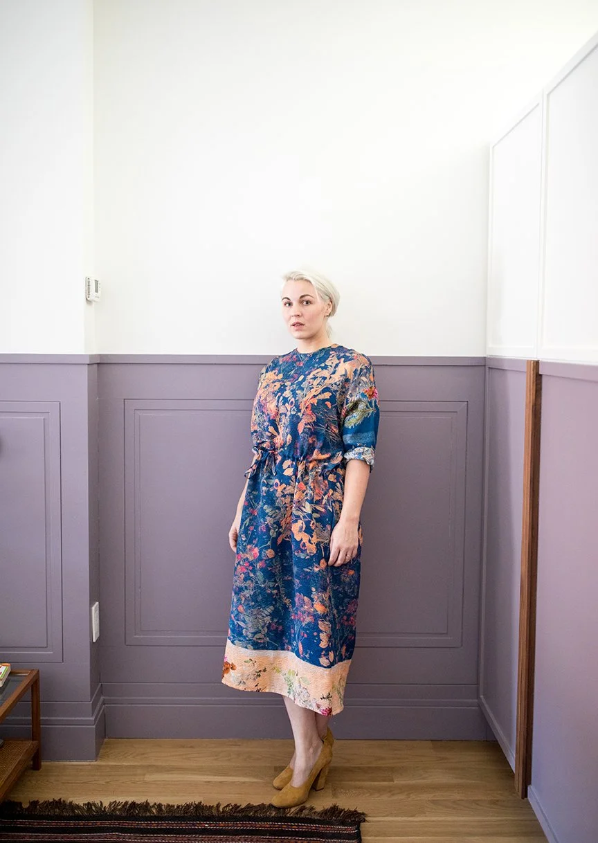 A woman with blonde hair wearing a colorful, patterned dress with a drawstring waist, standing in a corner of a room with purple wainscoting and wooden flooring.