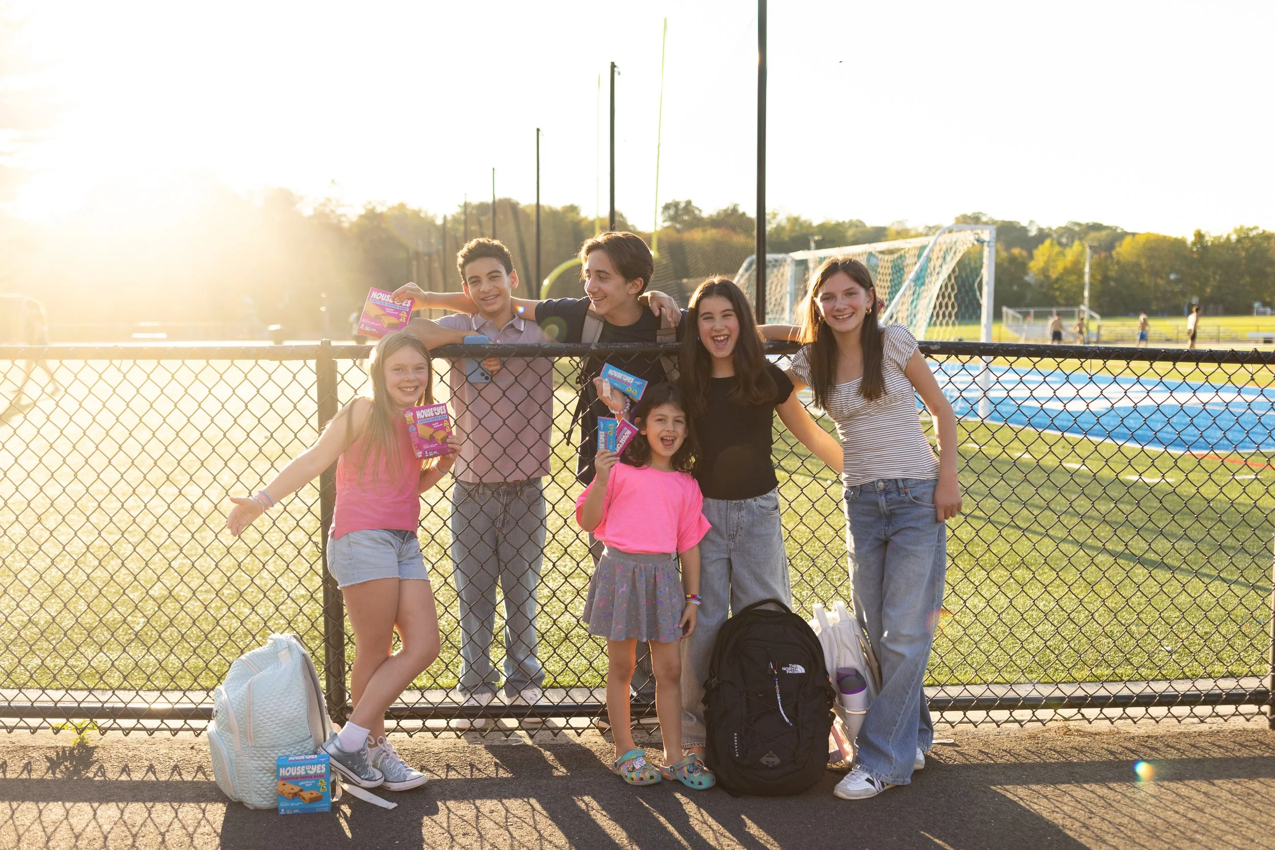 Group of children and teenagers at a sports field, standing behind a chain-link fence, smiling, with backpacks and snacks, during sunset.